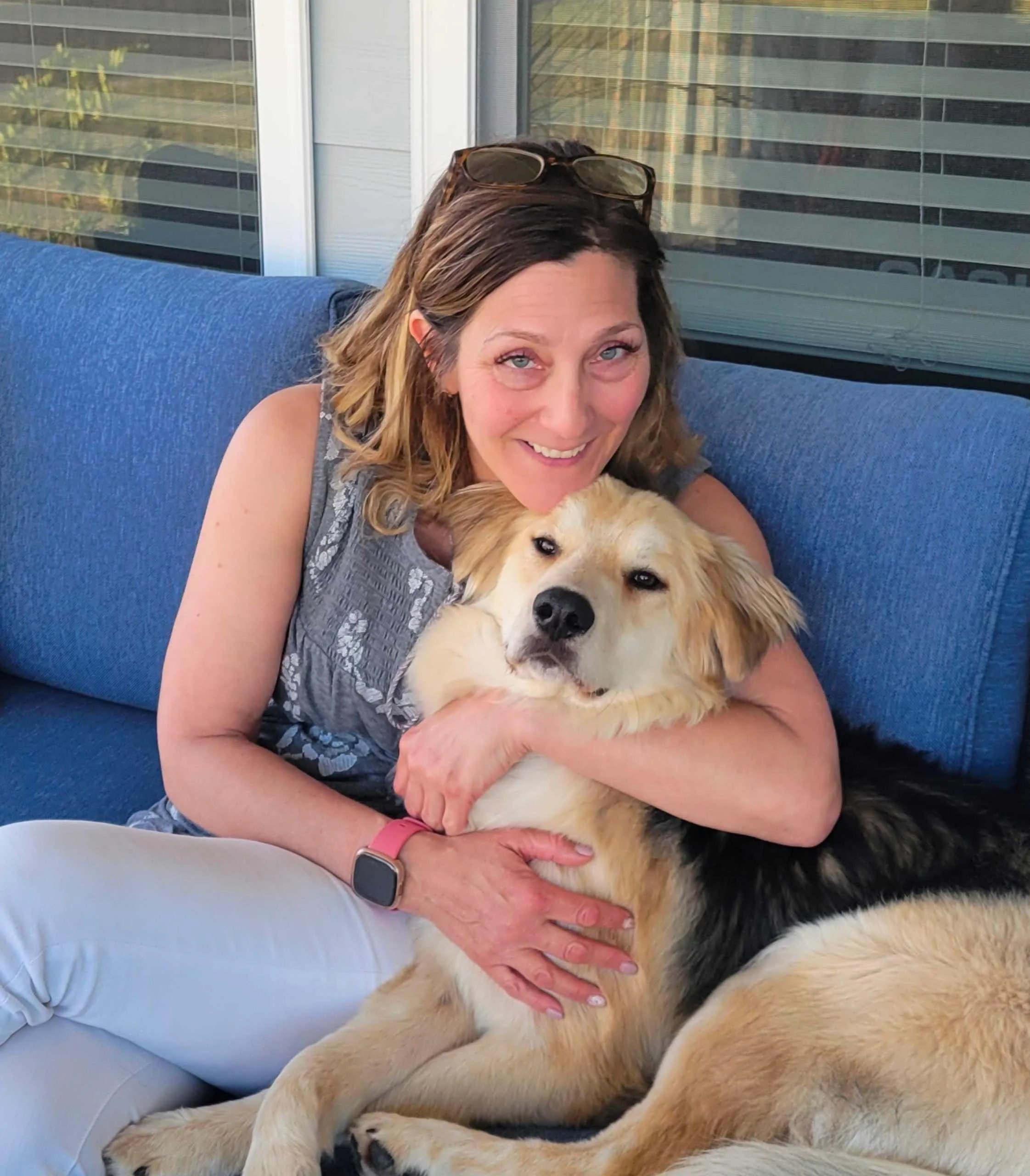 A woman with glasses resting on her head hugging a large, mixed-breed dog on a blue outdoor sofa in front of a glass window.