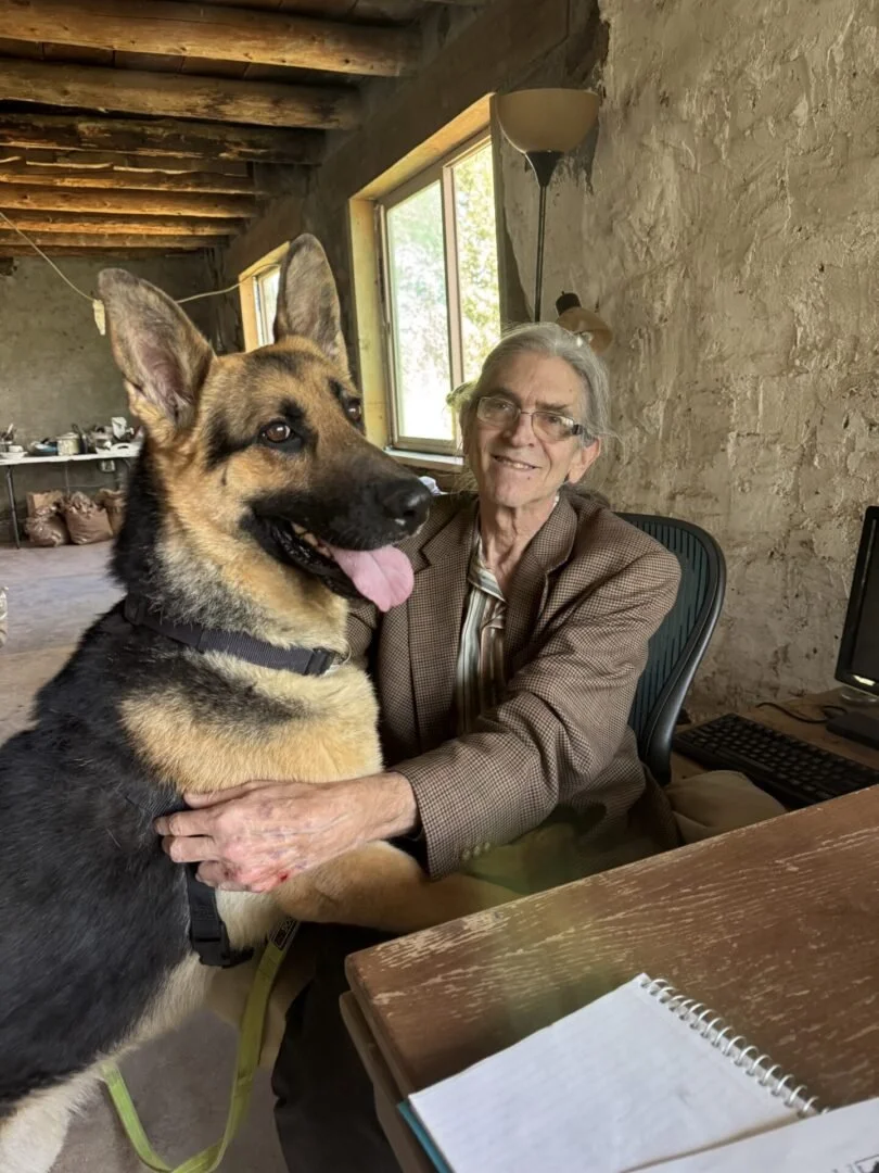 An elderly woman with glasses and gray hair smiling while hugging a large German Shepherd dog in a rustic room with a stone wall, a window, and a desk with a notebook and computer.