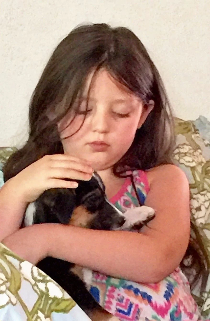 A young girl with long brown hair holding a small black and white puppy close to her chest with her eyes closed.