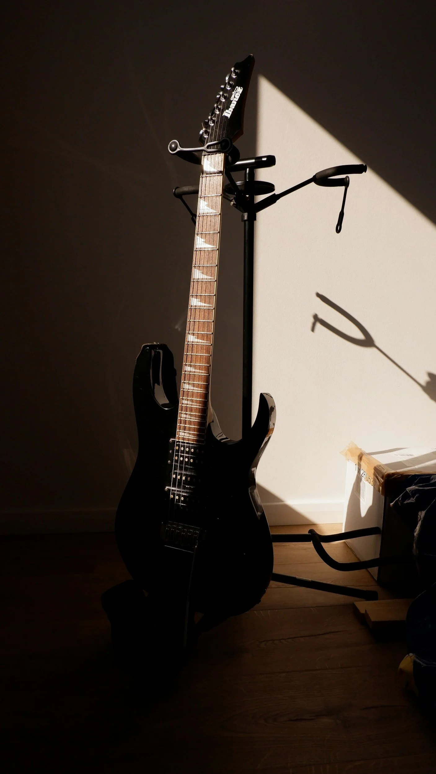 Black electric guitar leaning against a wall, casting a shadow, with sunlight illuminating part of the scene. There is a guitar stand and a cardboard box nearby.