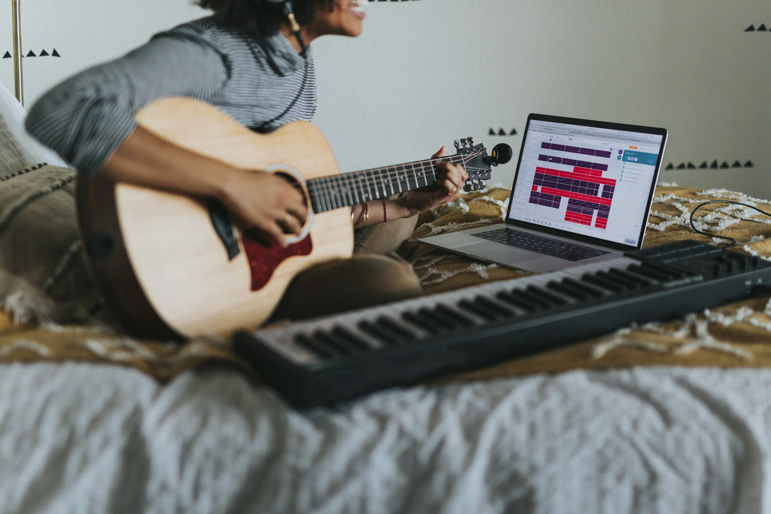 Person playing acoustic guitar on bed with a laptop displaying a digital audio workstation, a MIDI keyboard, and music editing software.