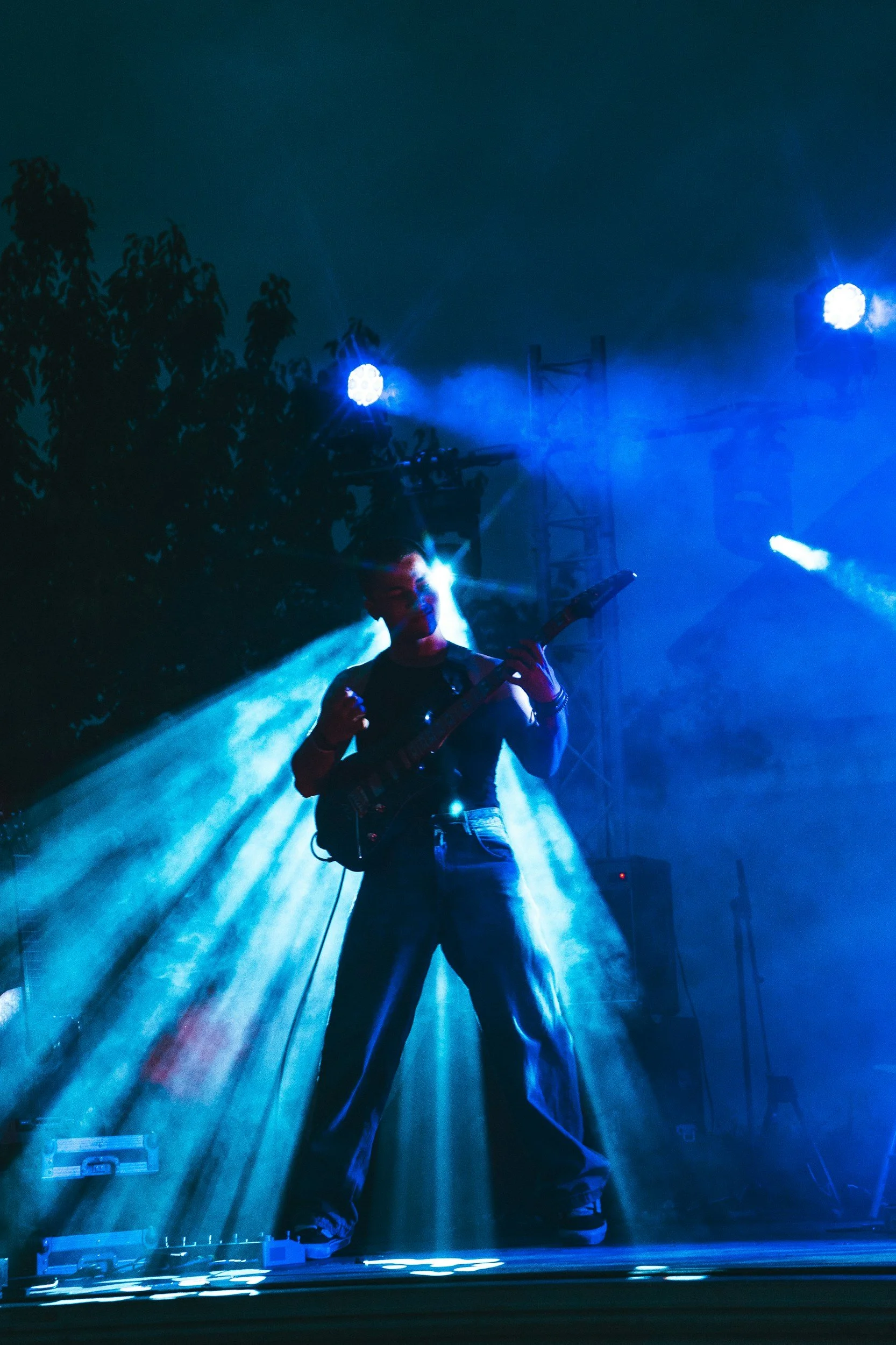 A musician playing an electric guitar on stage during a concert, with blue stage lights and fog creating a dramatic atmosphere.