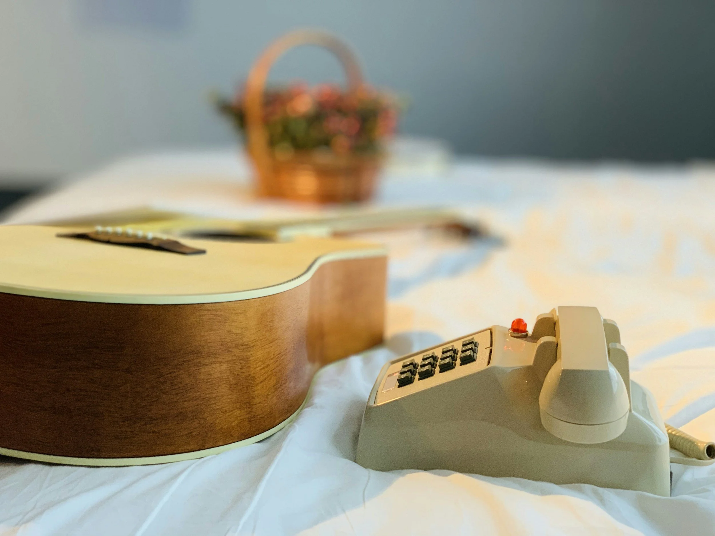 An electric guitar and an old rotary phone on a bed with a basket of flowers in the background.
