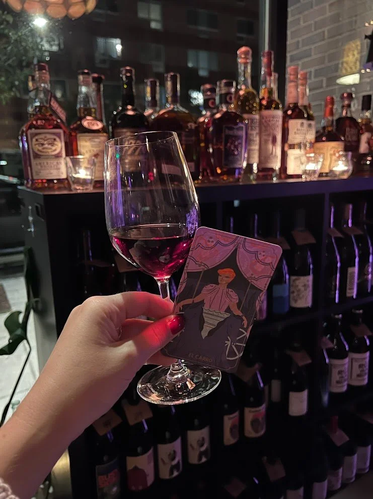 Person holding a glass of red wine and a tarot card in front of a bar with various bottles of liquor on shelves behind.