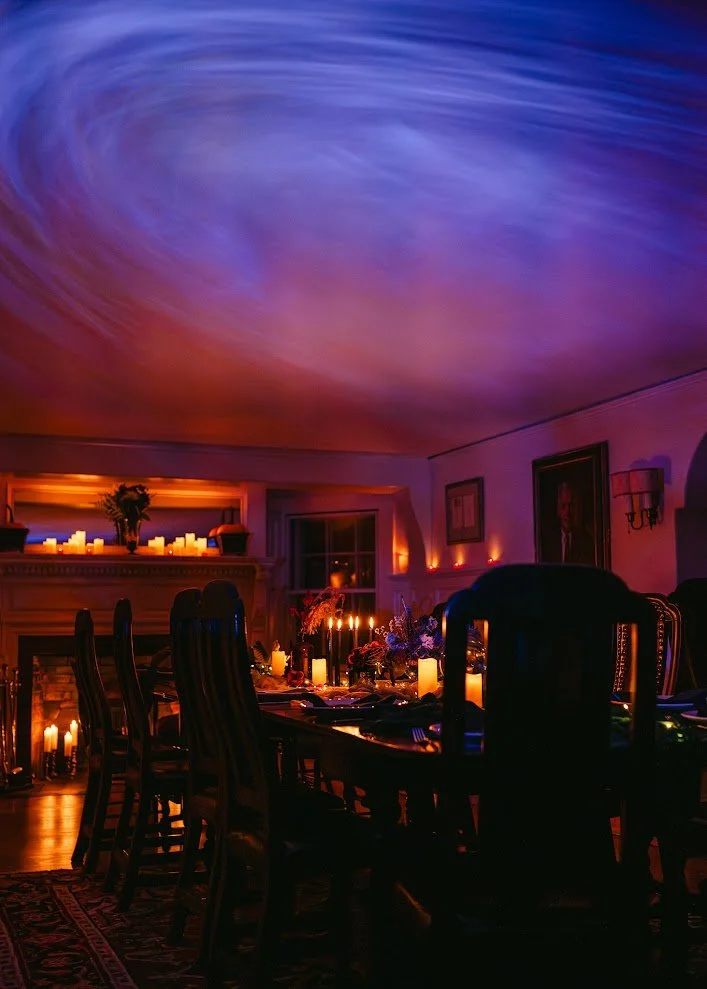 A dining room with candles and dim lighting, illuminated by a colorful ceiling projection resembling a swirling galaxy.
