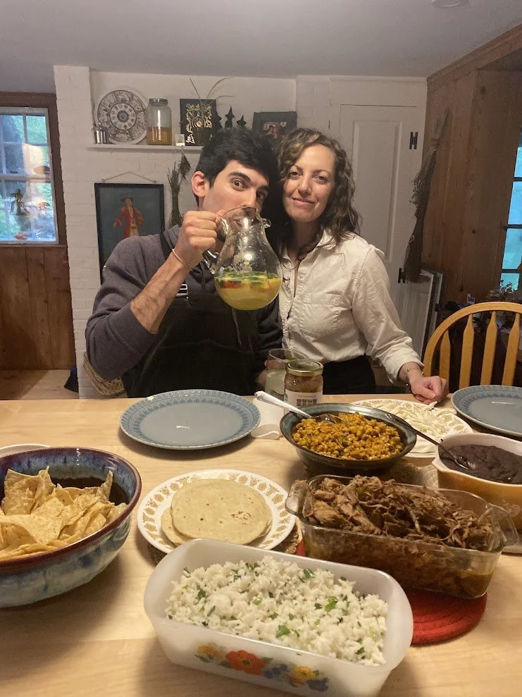 Two people sitting at a dining table with various dishes, one holding a glass pitcher of yellow beverage, in a cozy home interior.