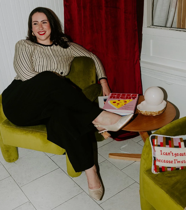 A woman sitting on a green velvet sofa, smiling, with her legs crossed on a small round wooden table. She has dark hair, red lipstick, and is wearing a striped long-sleeve top, black pants, and beige heels. The table holds a pizza magazine and a spherical white decorative object. There's a pillow with red and black text and a colorful pattern on a green chair nearby, and a red curtain with a window behind her.