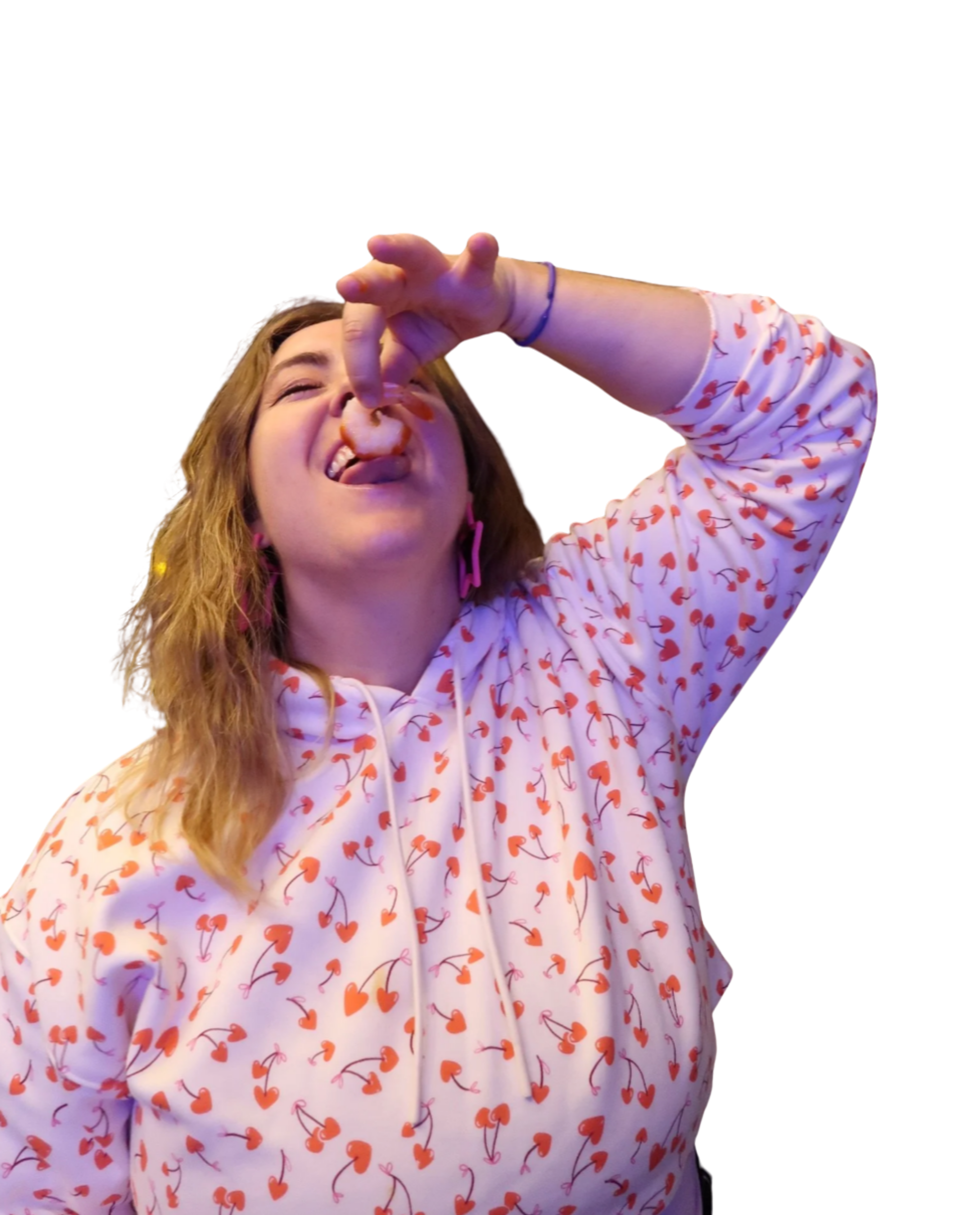 A young woman with wavy reddish-brown hair, wearing a white hoodie with red cherry print, making a silly face with her tongue out and her eyes squinting, holding a shrimp near her mouth.