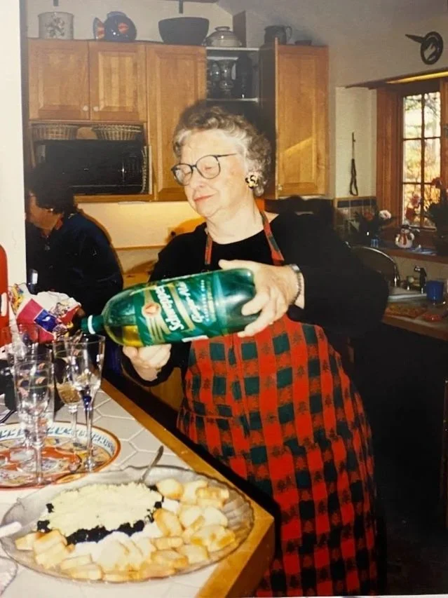An elderly woman with glasses and earrings, wearing a red and black checkered apron, pouring a green bottle of sparkling water into glasses in a kitchen. There are dishes, glasses, and a decorated plate of food on the counter.