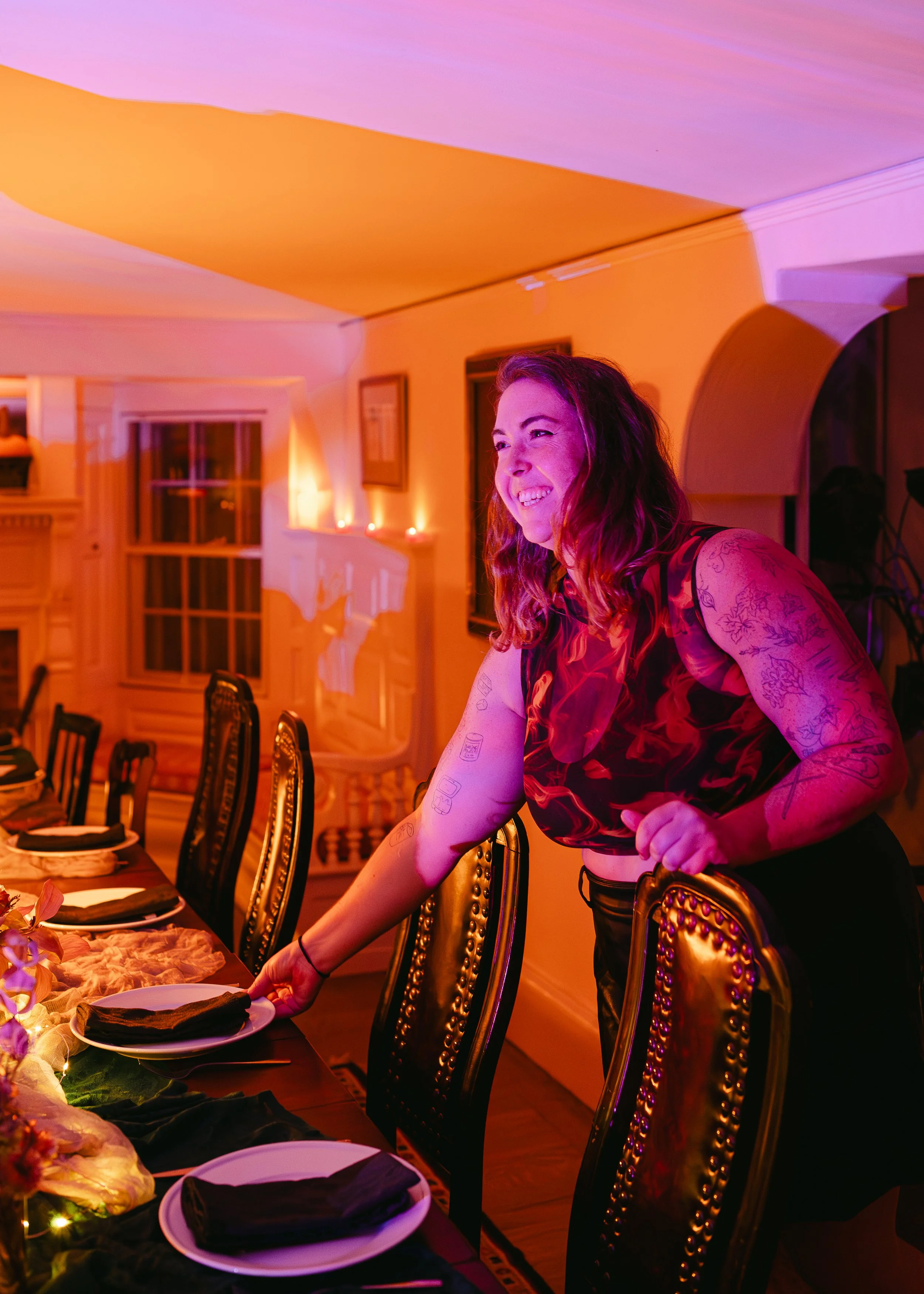 Woman smiling while setting a dinner table with black napkins in a warmly lit room with elegant chairs and holiday decor.