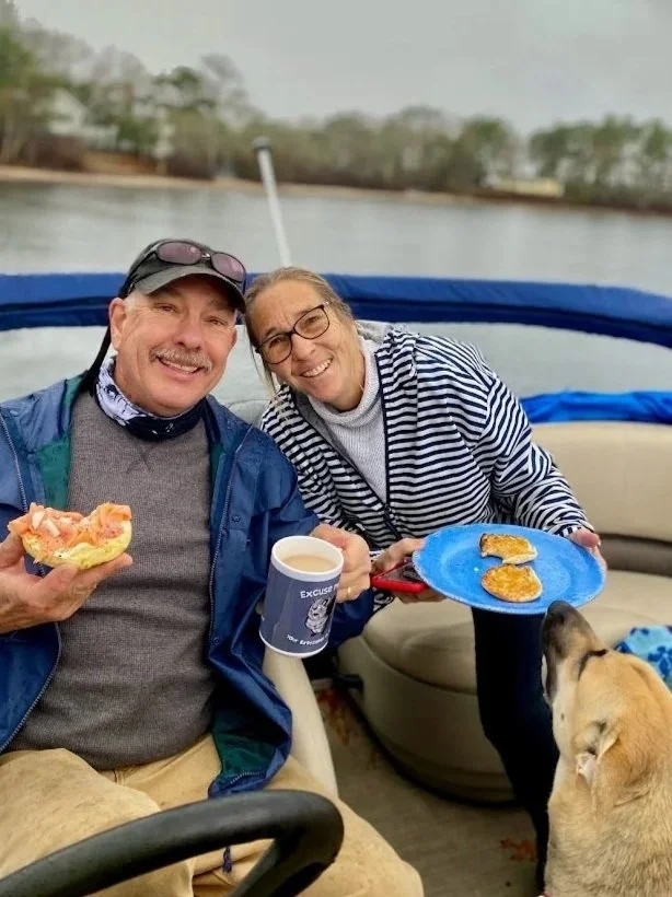 A smiling man and woman on a boat, holding breakfast food and coffee cups, with a dog looking up at the food, and a river in the background.