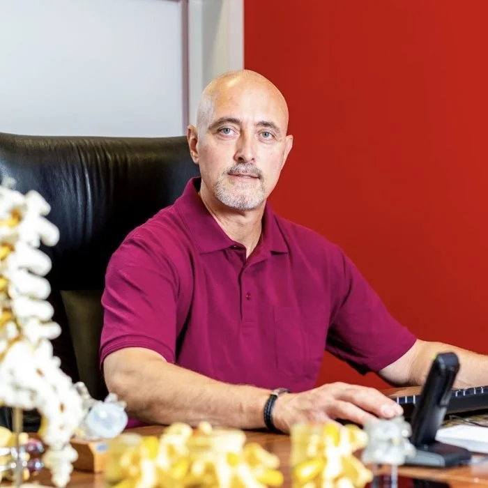 Mann sitzt an einem Schreibtisch vor einer roten Wand, trägt ein dunkleres rotes Hemd, hat einen Laptop und Dekorationen auf dem Tisch.