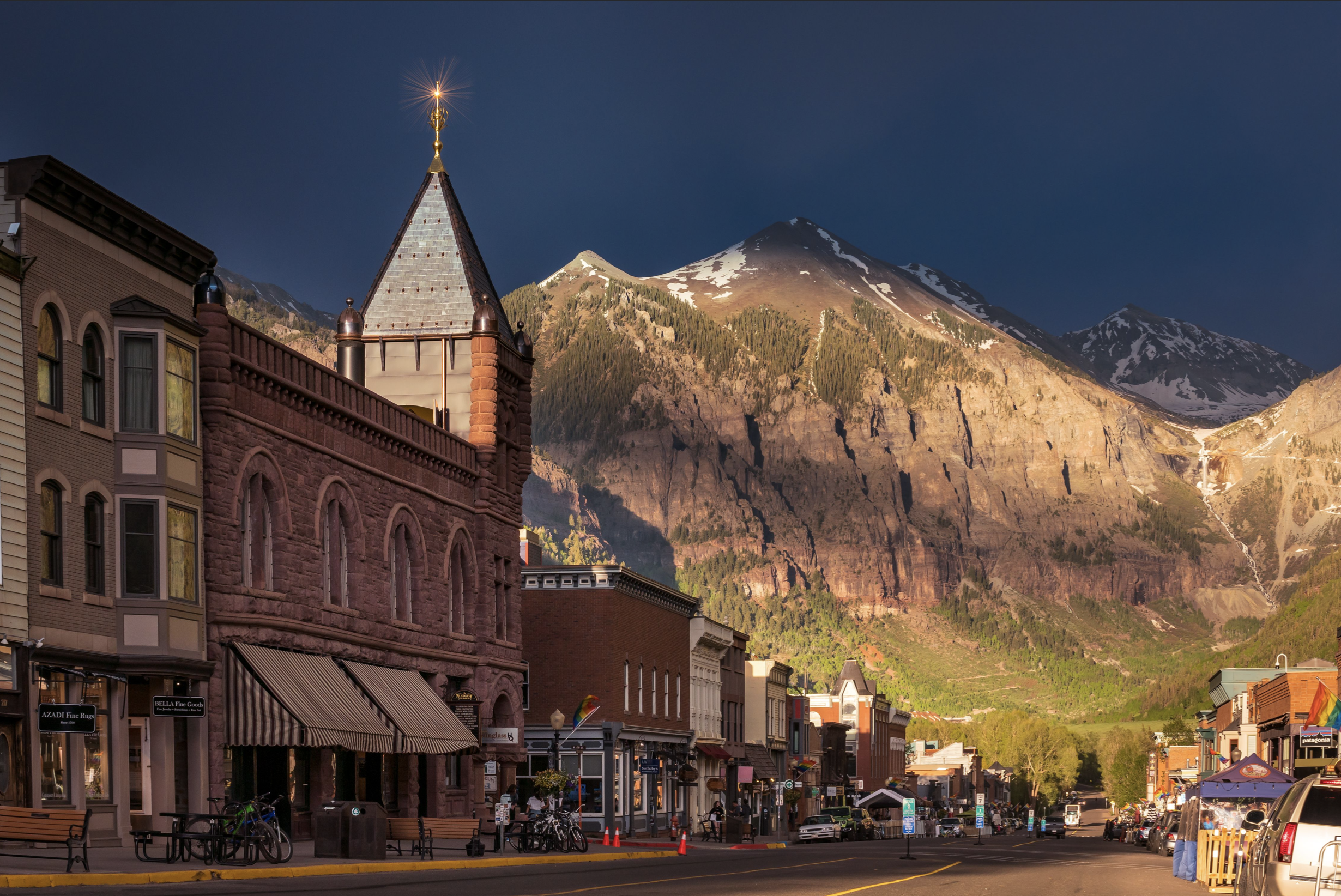 A small mountain town street with historic buildings and shops, set against tall mountains with snow on their peaks at sunset.