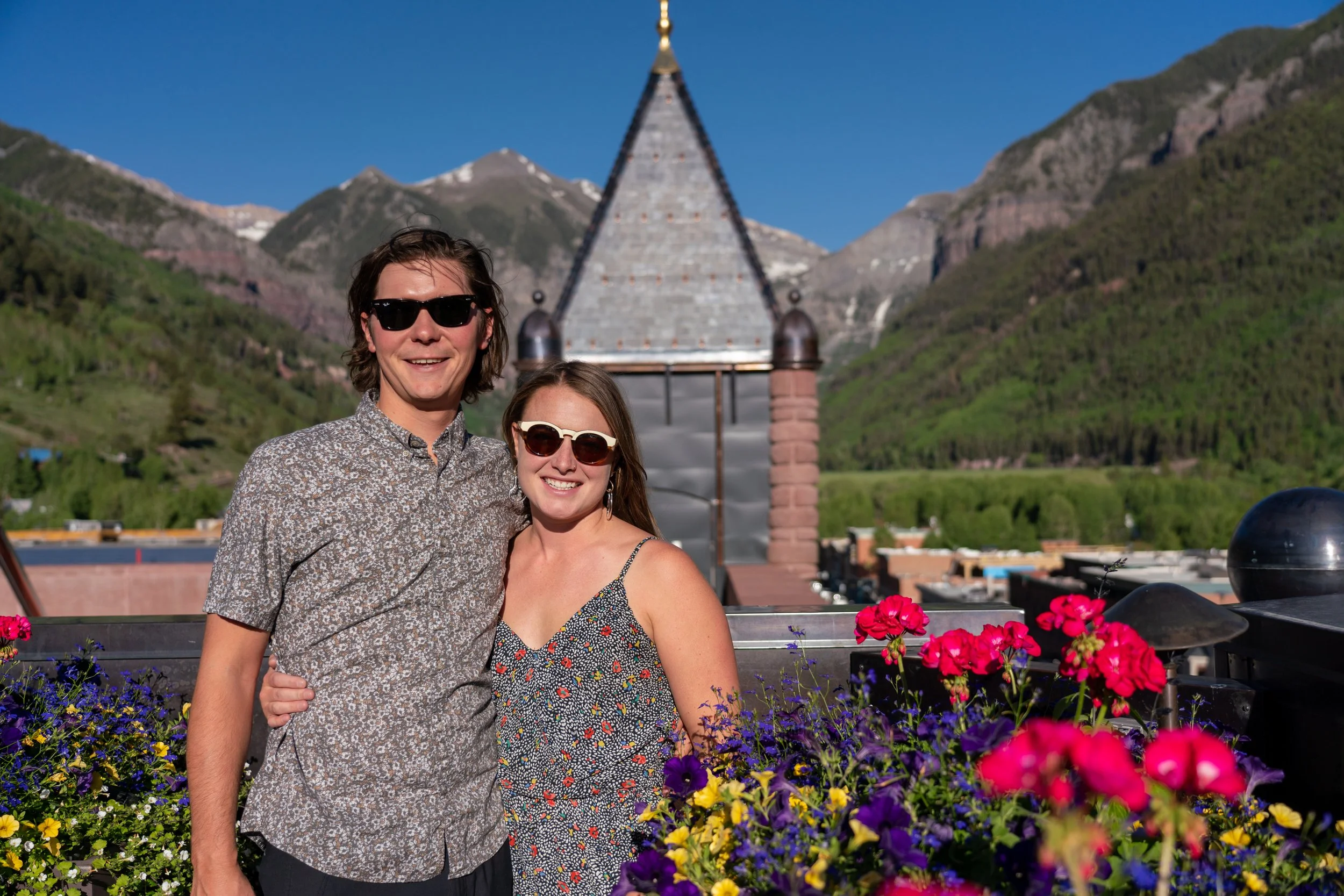 A smiling young man and woman wearing sunglasses standing close together on a rooftop terrace with colorful flowers. Behind them are mountains and a building with a steep, pointed roof.