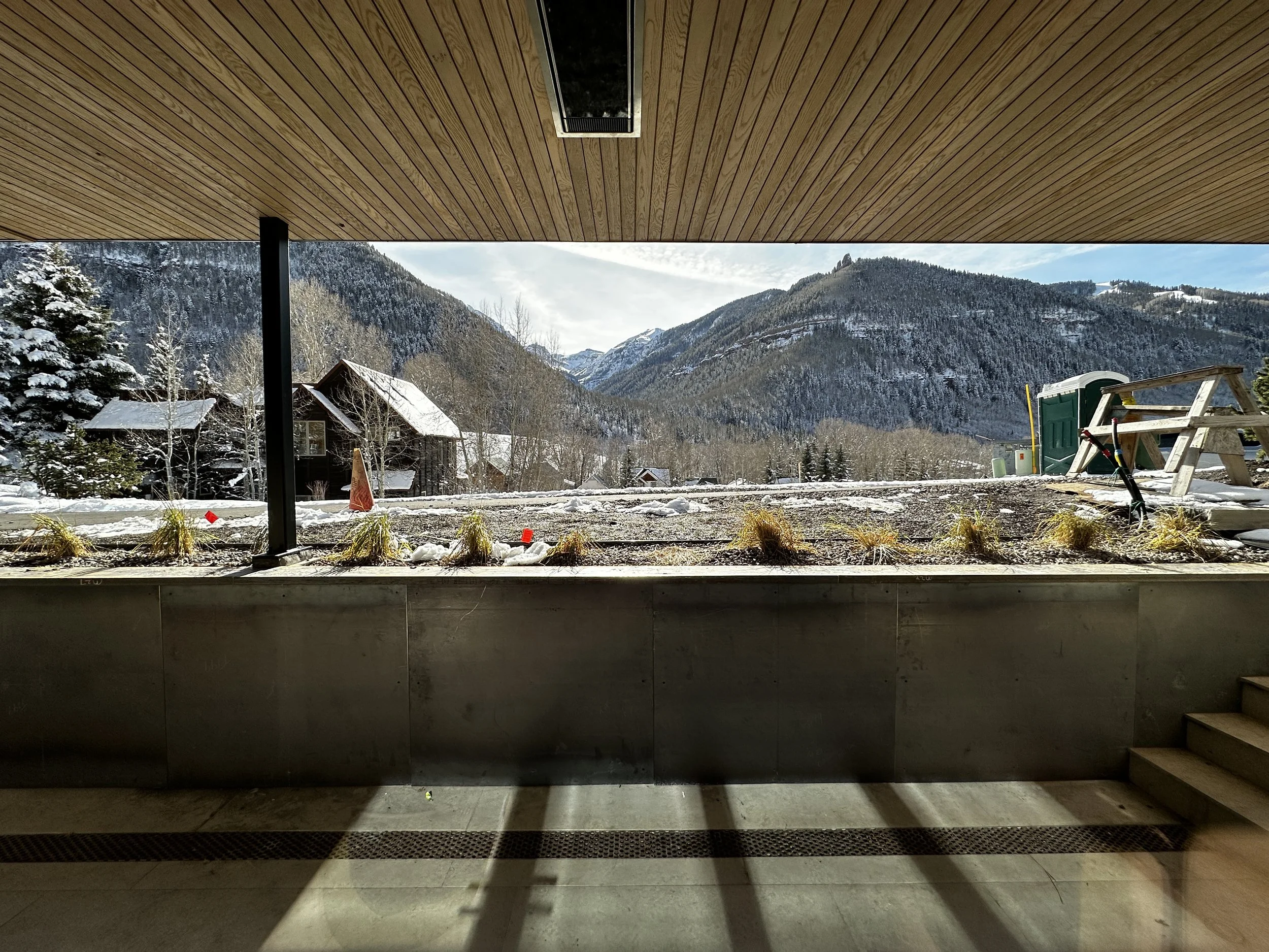 View from inside a building with a large open window, revealing snow-covered mountains, trees, and houses outside, during winter.