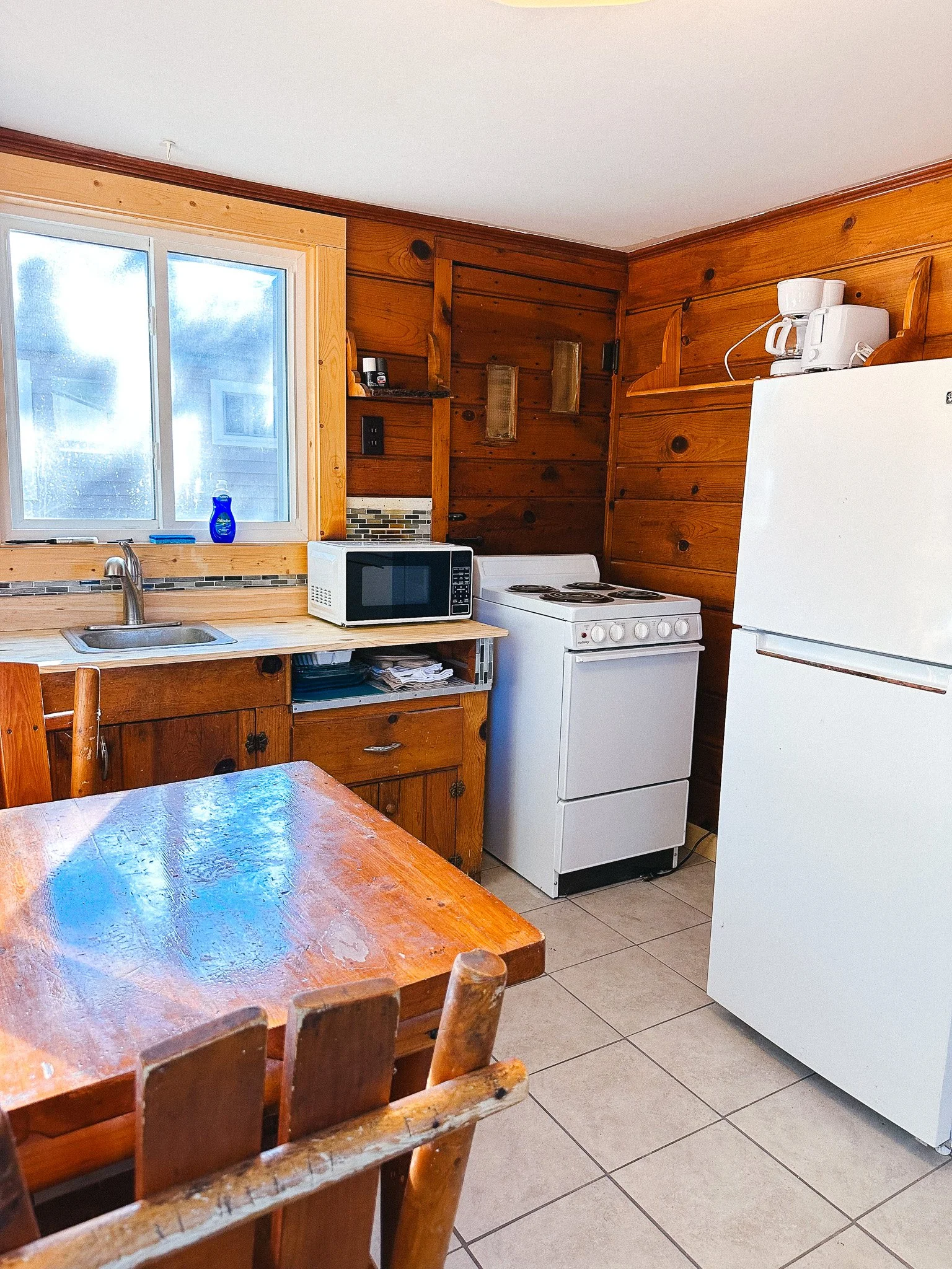 A rustic kitchen with wooden walls and cabinetry, a window over the sink, a microwave on the counter, a white stove, and a white refrigerator. There is also a wooden dining table and chairs.