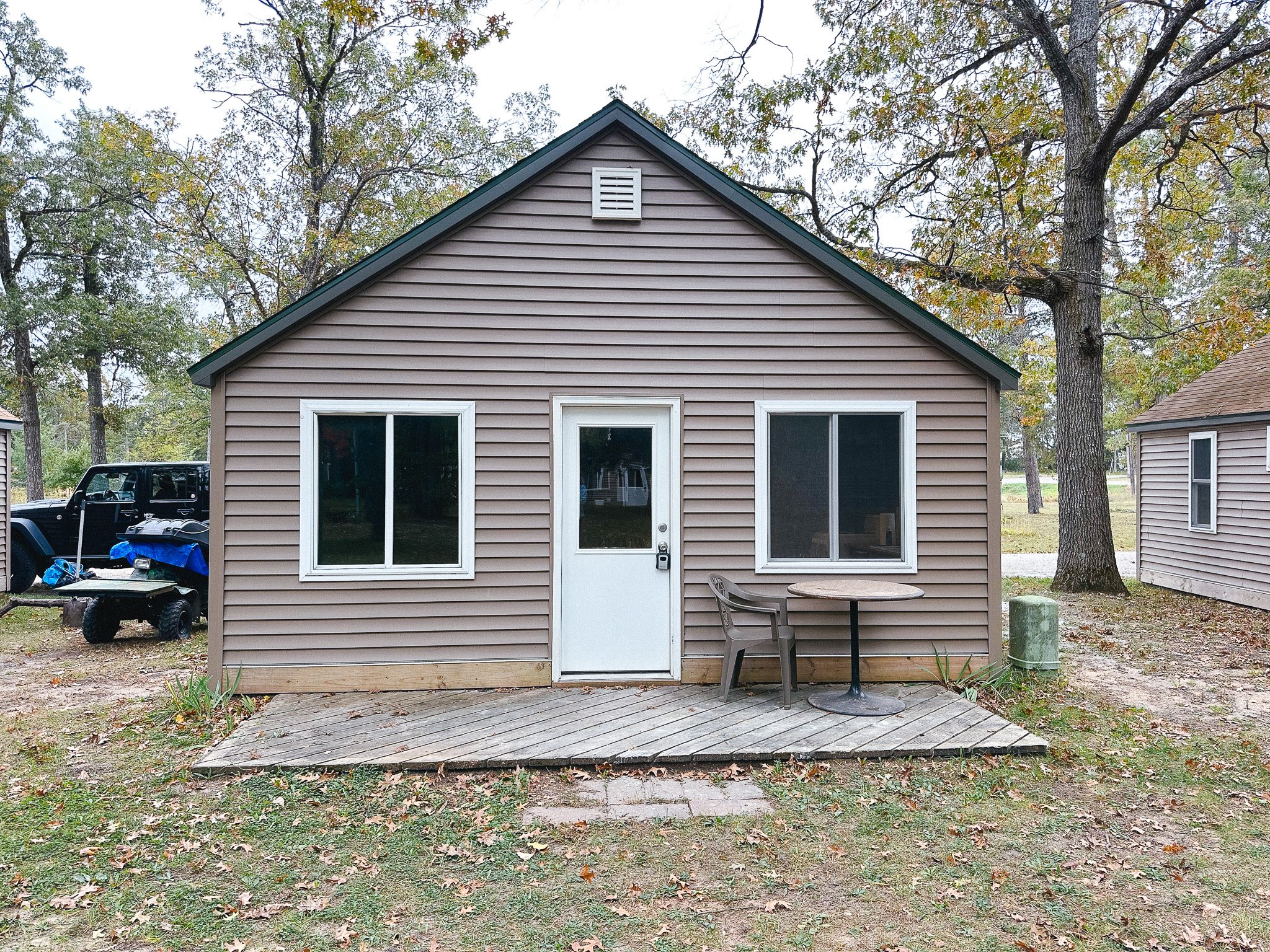 Small house with beige siding, white door, and two windows. Small wooden deck with a plastic chair and round table outside. Surrounding yard has trees, grass, and leaves, with a black utility vehicle and piles of items on the left.