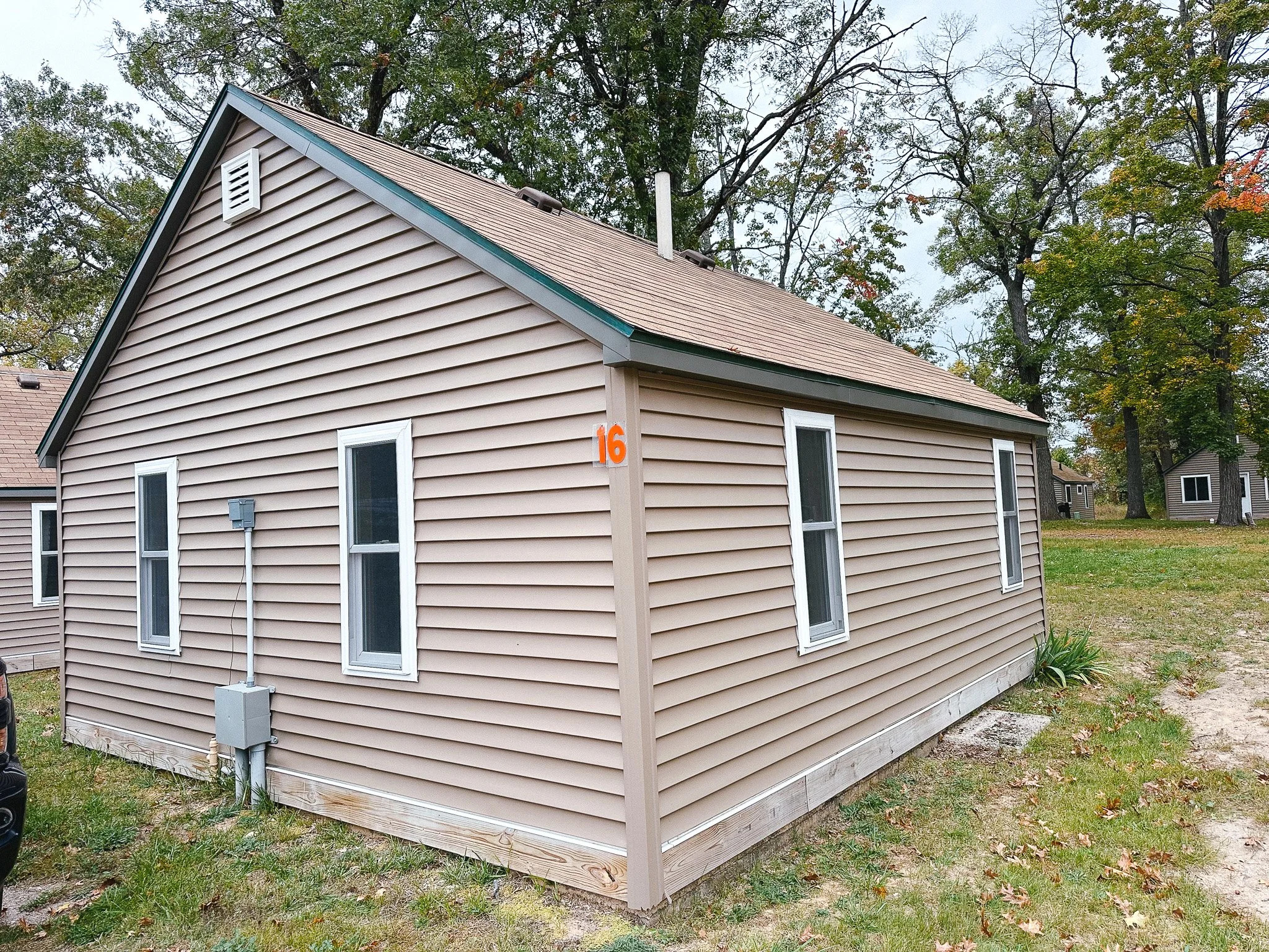 Side view of a beige house with white-framed windows and a sloped roof, numbered 16, surrounded by trees and grass.