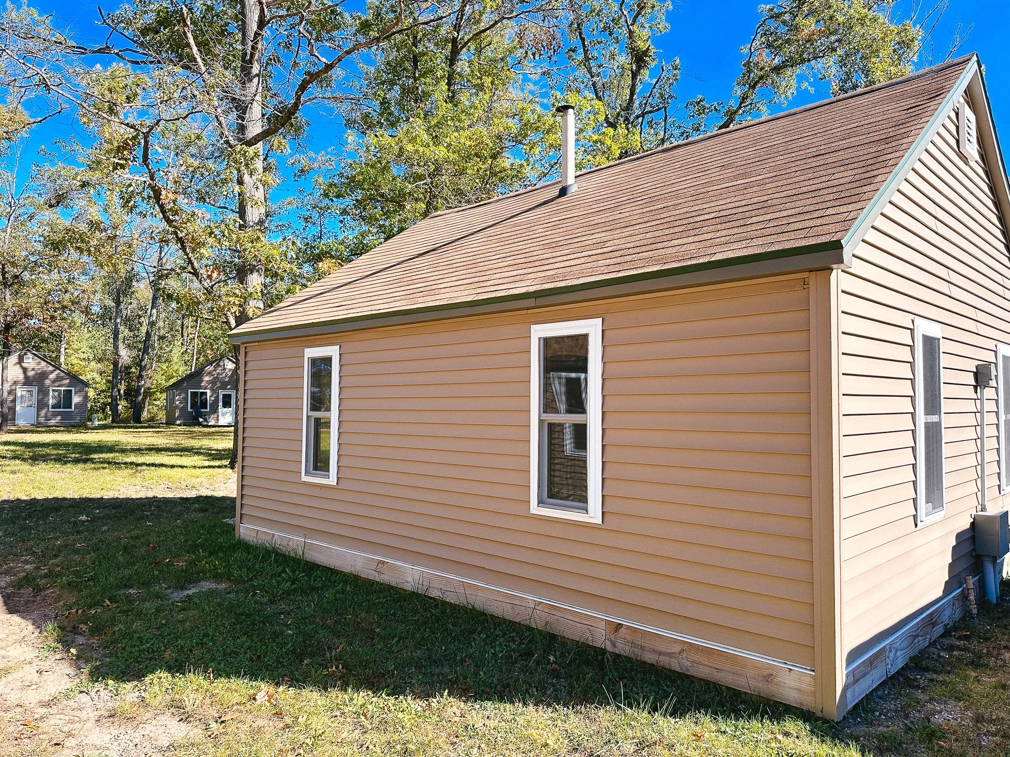A beige house with white-trimmed windows and a brown shingle roof, surrounded by green grass and trees under a blue sky.