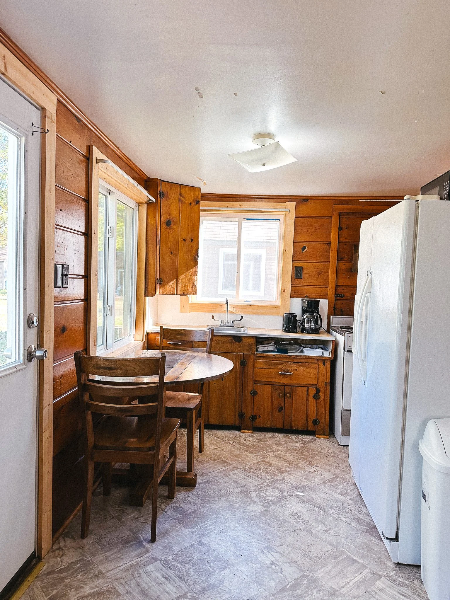 Cozy kitchen with wooden walls, a small round dining table with two chairs, a window above the sink, and various appliances including coffee maker, toaster, and refrigerator.