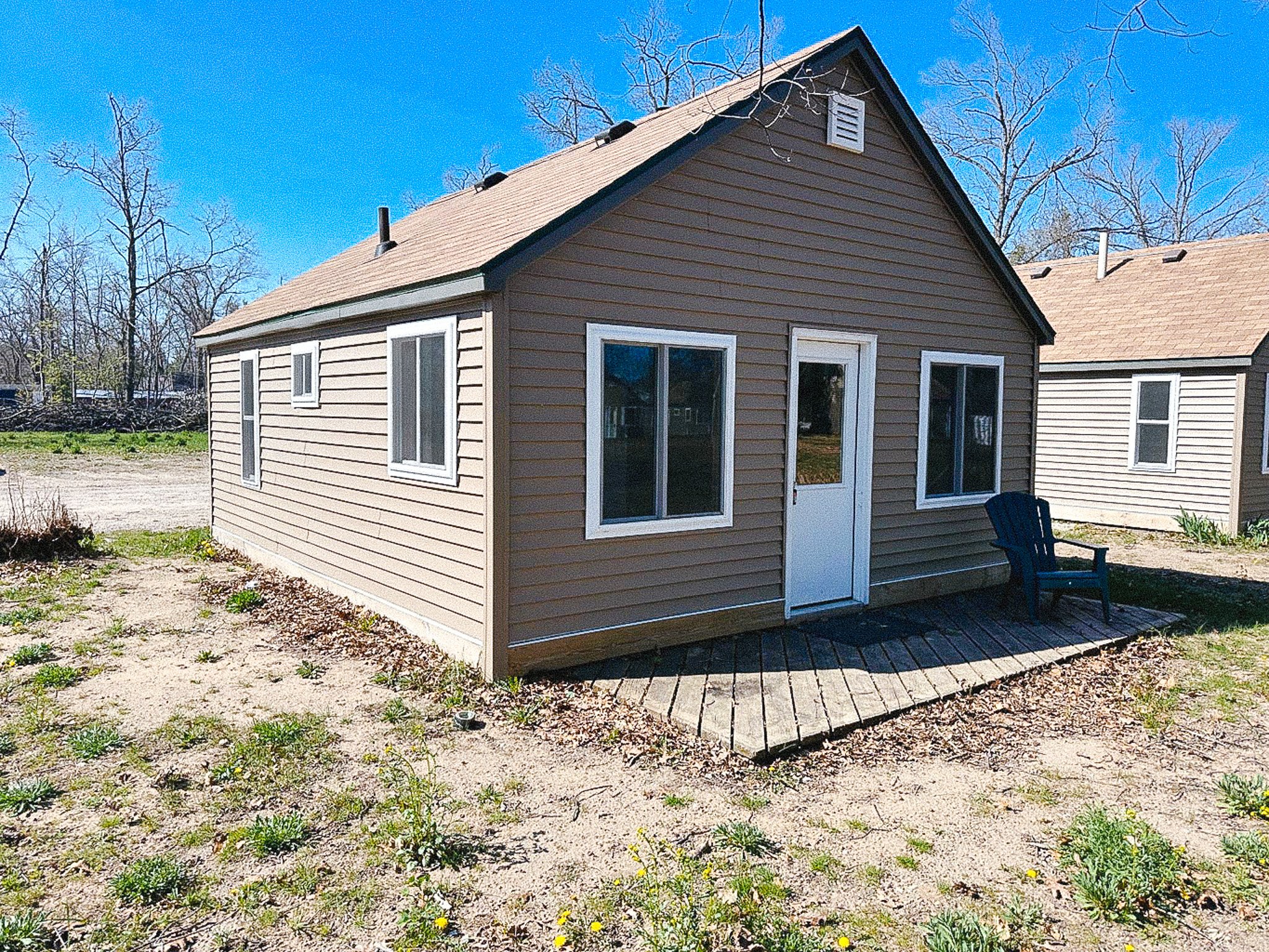 A small single-story house with tan siding, white trim, and a gable roof. It has three windows and a glass door leading to a small wooden porch with a black plastic chair on it. The yard is bare with some sparse grass, and the sky is clear and blue.