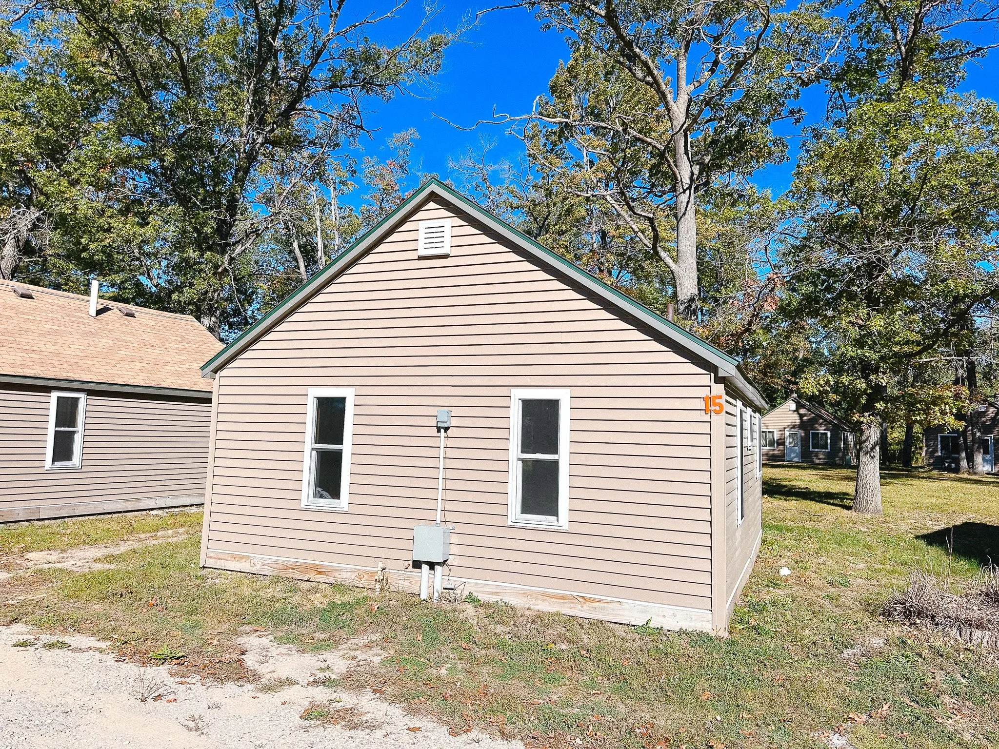 A small beige house with white trim, three windows, and a green roof, situated in a grassy yard with trees in the background.
