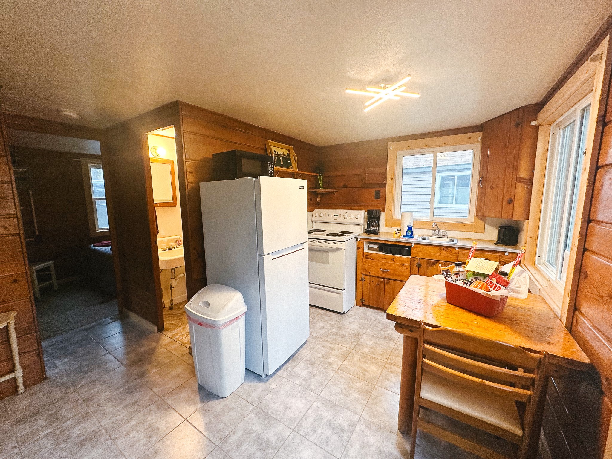A rustic kitchen with wooden cabinets and tiled flooring, featuring a white refrigerator, a stove, a microwave on top of the refrigerator, a coffee maker, sink, and a small dining table with chairs, with large windows letting in natural light.