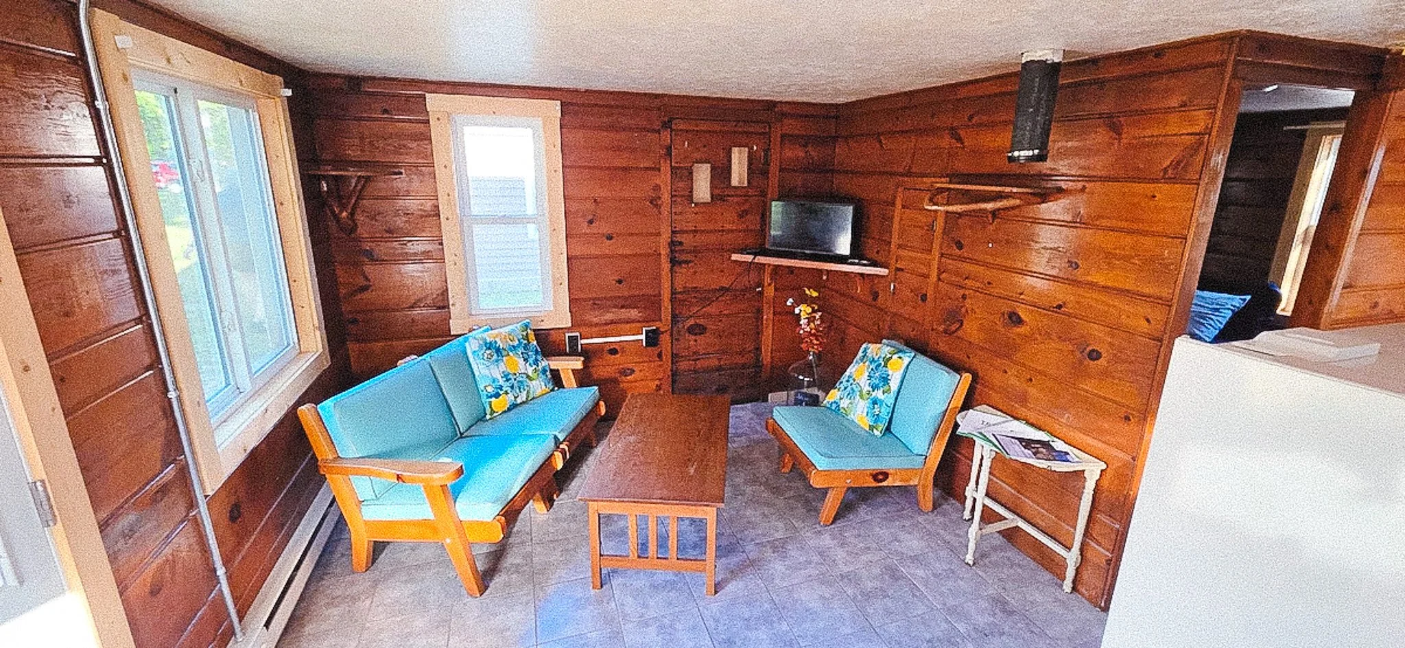 Living room with wood-paneled walls, featuring two blue sofas with floral cushions, a coffee table, a small side table with magazines, a wall-mounted TV, a window, and a doorway leading to a bedroom.