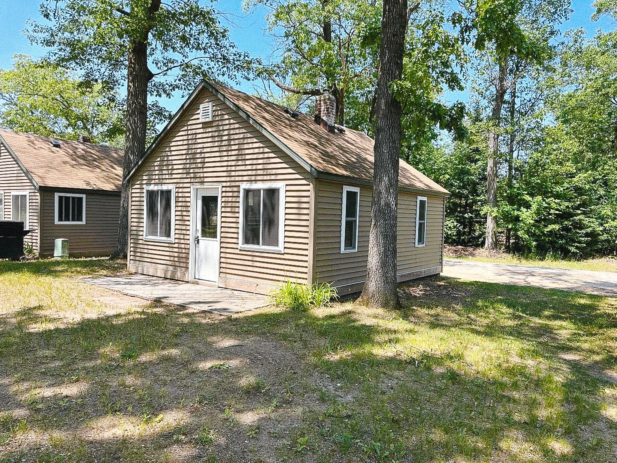 A small beige house with white trim surrounded by trees and tall grass on a sunny day.