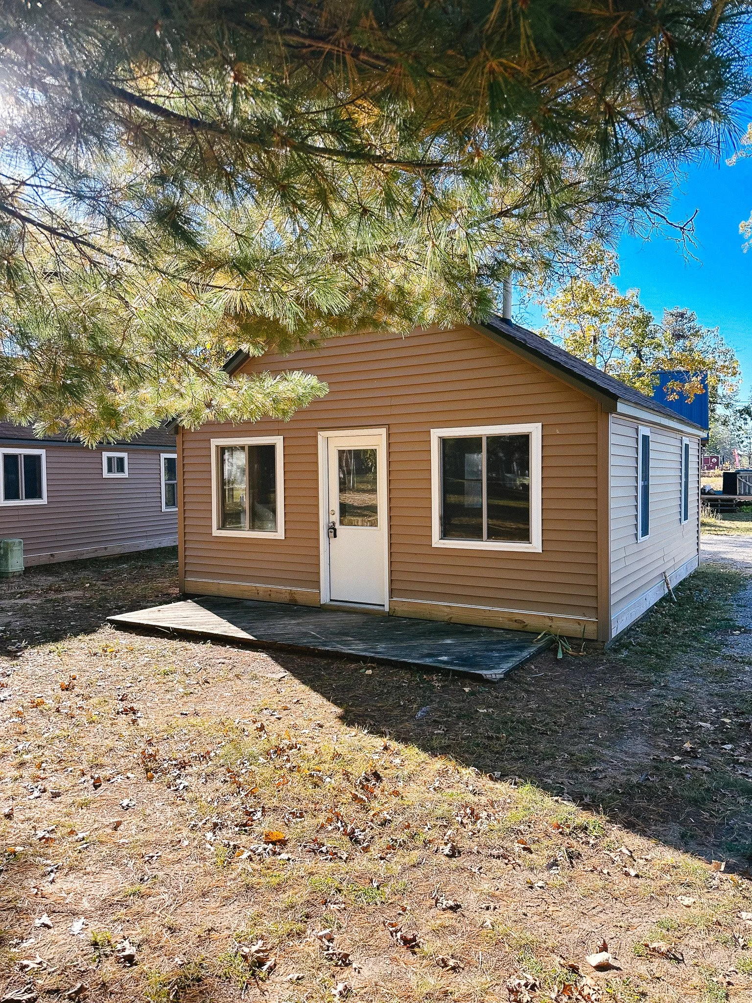 A small house with tan siding, a white door, and two windows on the front, with a small wooden porch. There are trees and blue sky in the background.
