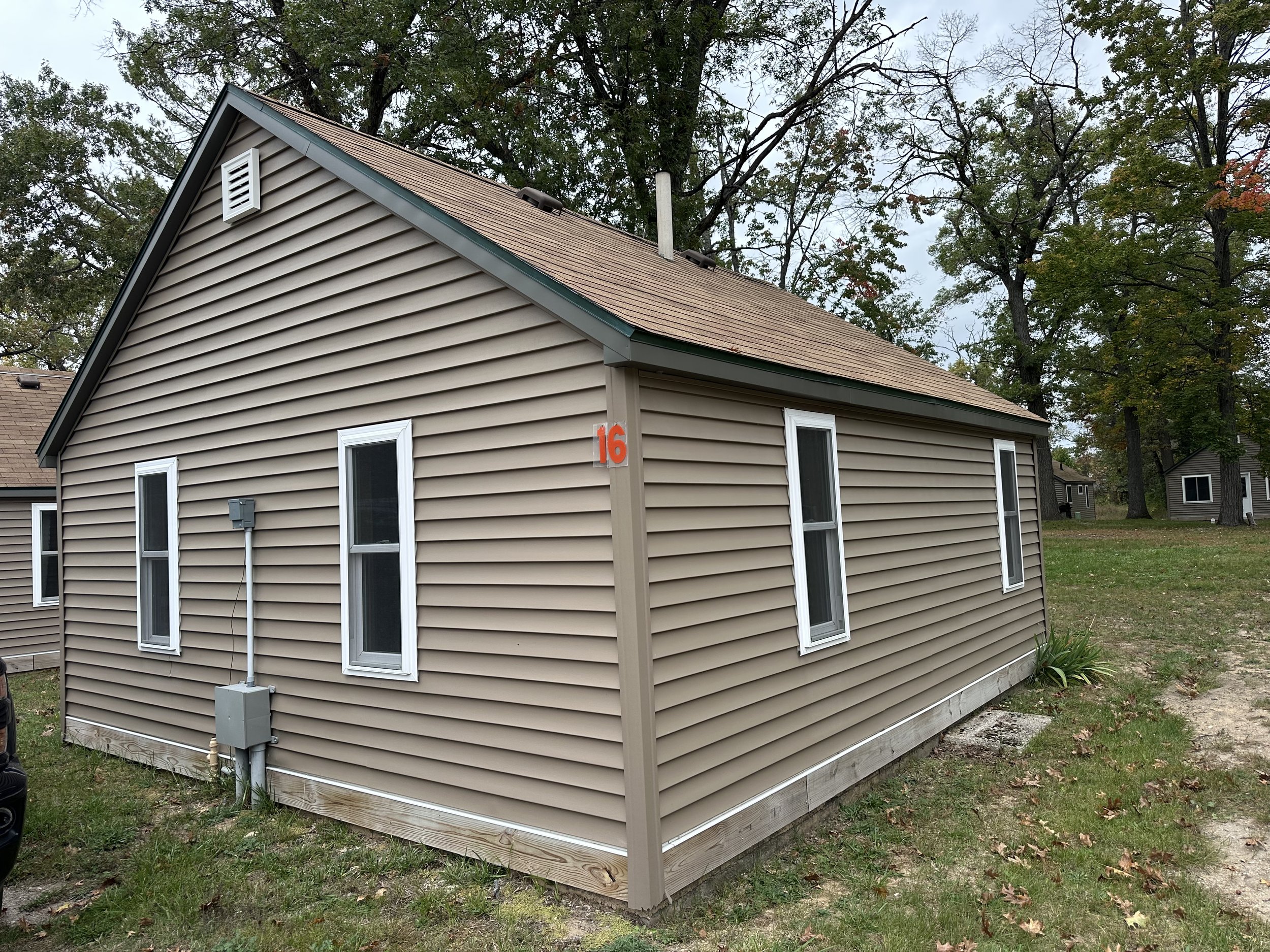 Side view of a beige house with four windows, a brown roof, a gray electrical box, and a yard with grass and fallen leaves.