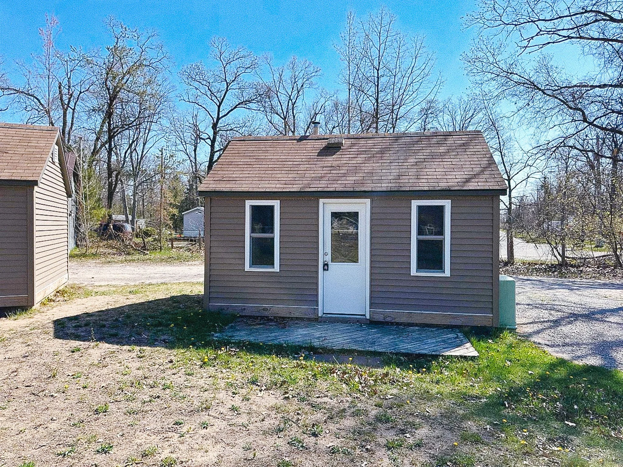 Small brown shed with a white door and two windows, surrounded by leafless trees on a sunny day.