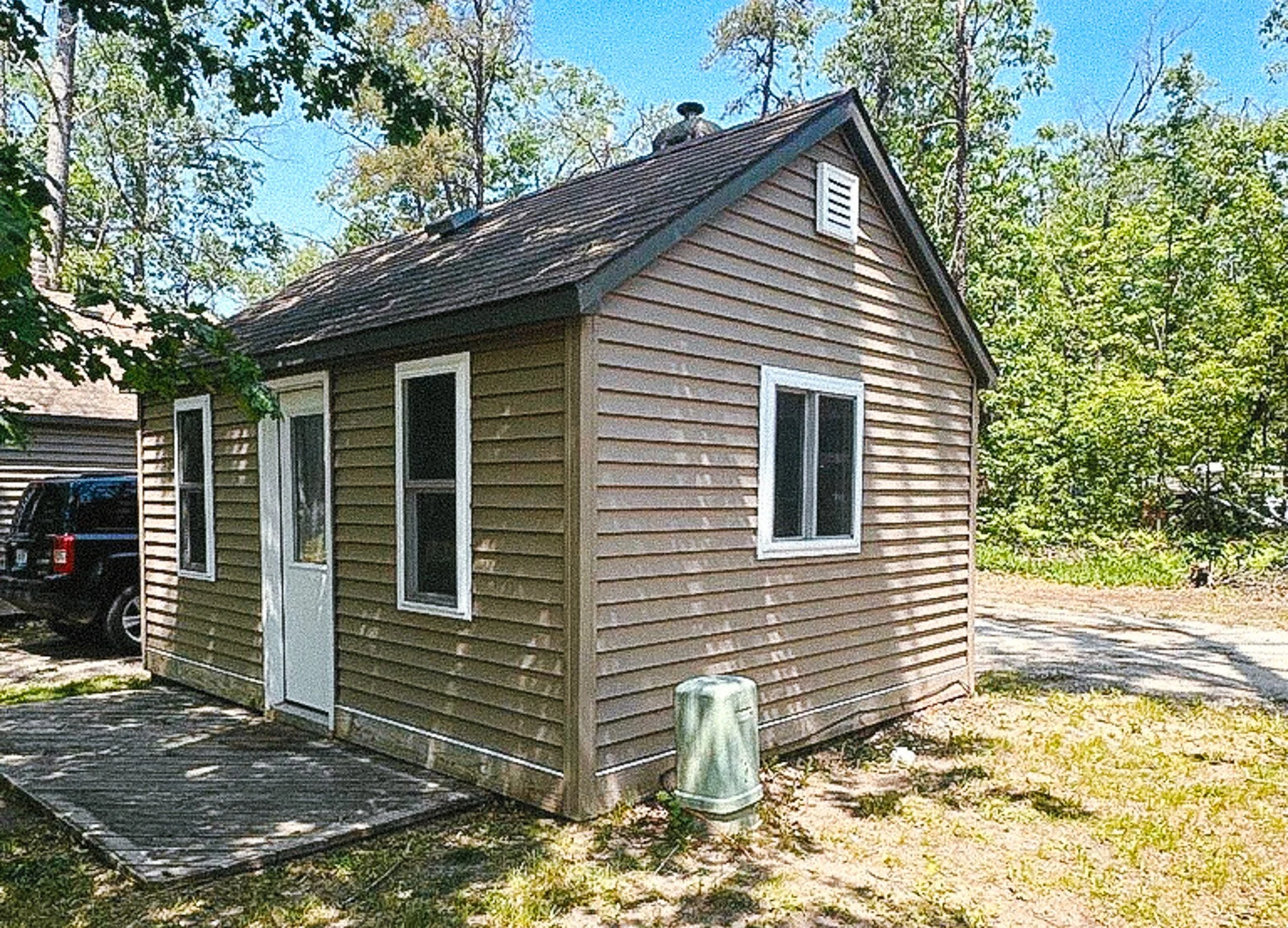 A small wooden house with beige horizontal siding, three windows, a white door, and a pitched roof with a ventilation louver and a chimney pipe, surrounded by trees and a gravel driveway.