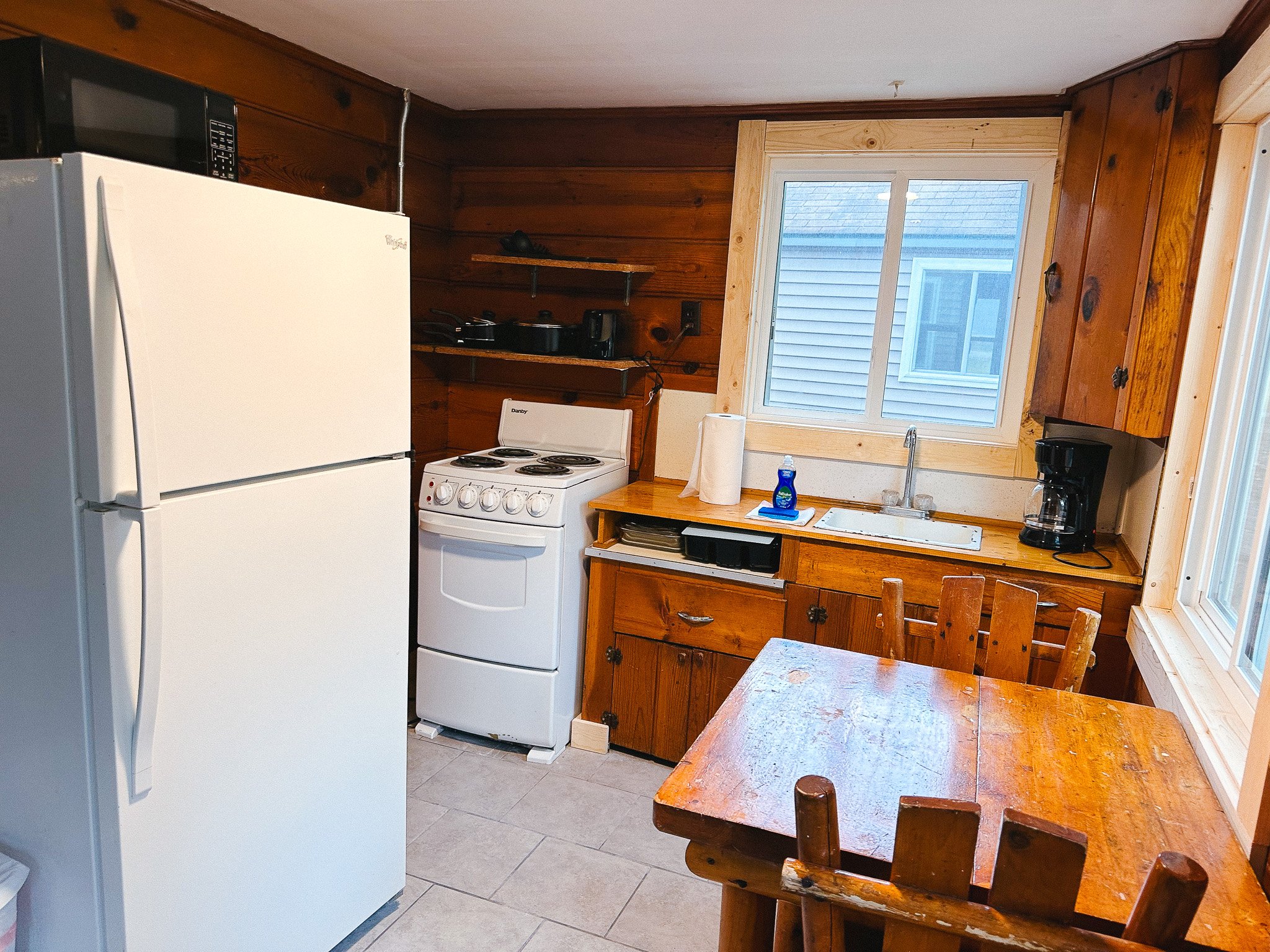 A rustic kitchen with wood-paneled walls, a white refrigerator, a small white stove, a wooden table with chairs, a window above the sink, and kitchen essentials like a coffee maker, paper towels, and dish soap.