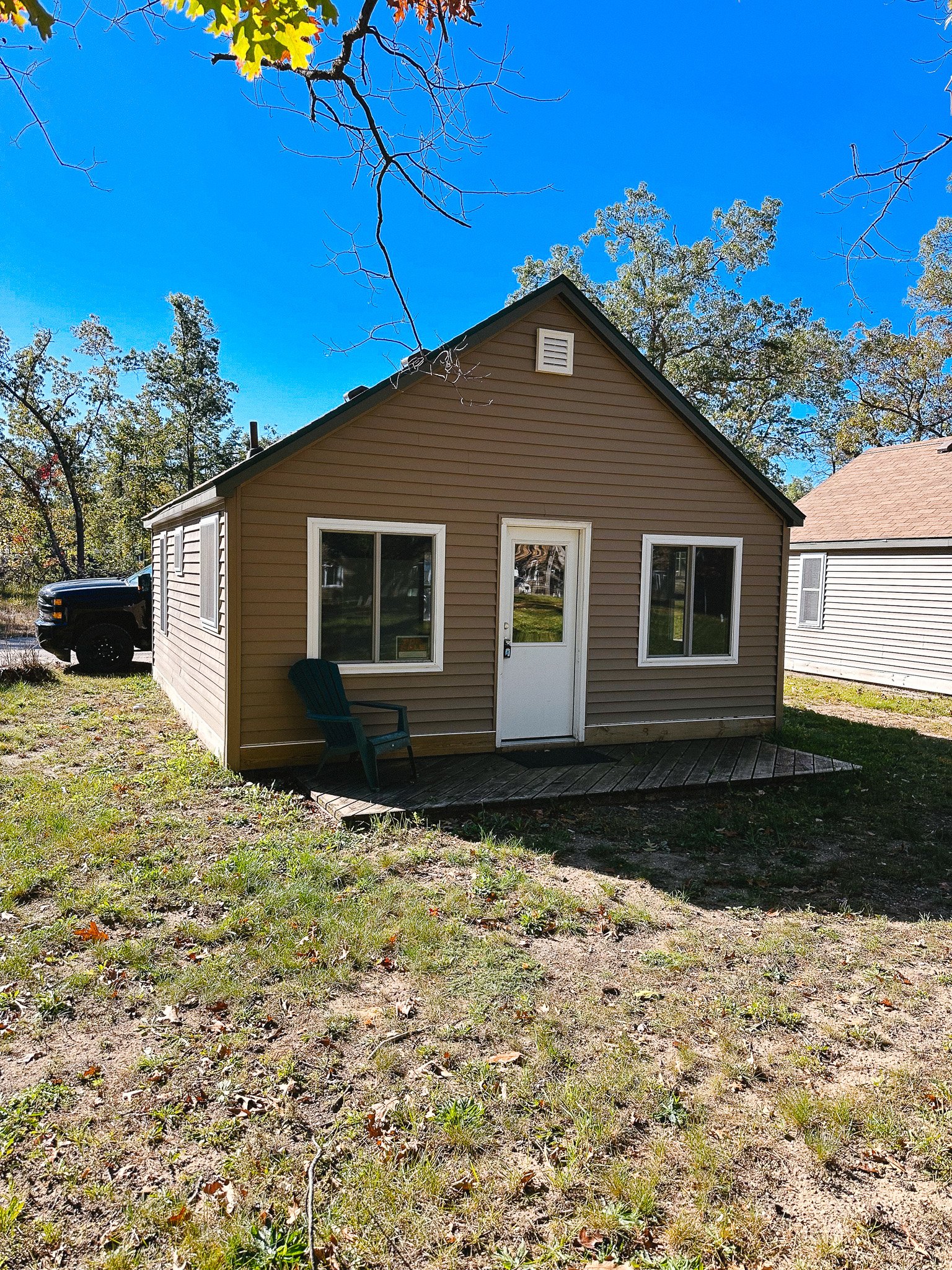 Small brown house with three windows, a white door, and a small wooden deck. A green chair sits on the deck. The house is surrounded by a grassy yard, and a black truck is parked to the left. The sky is clear and blue with some trees in the background.