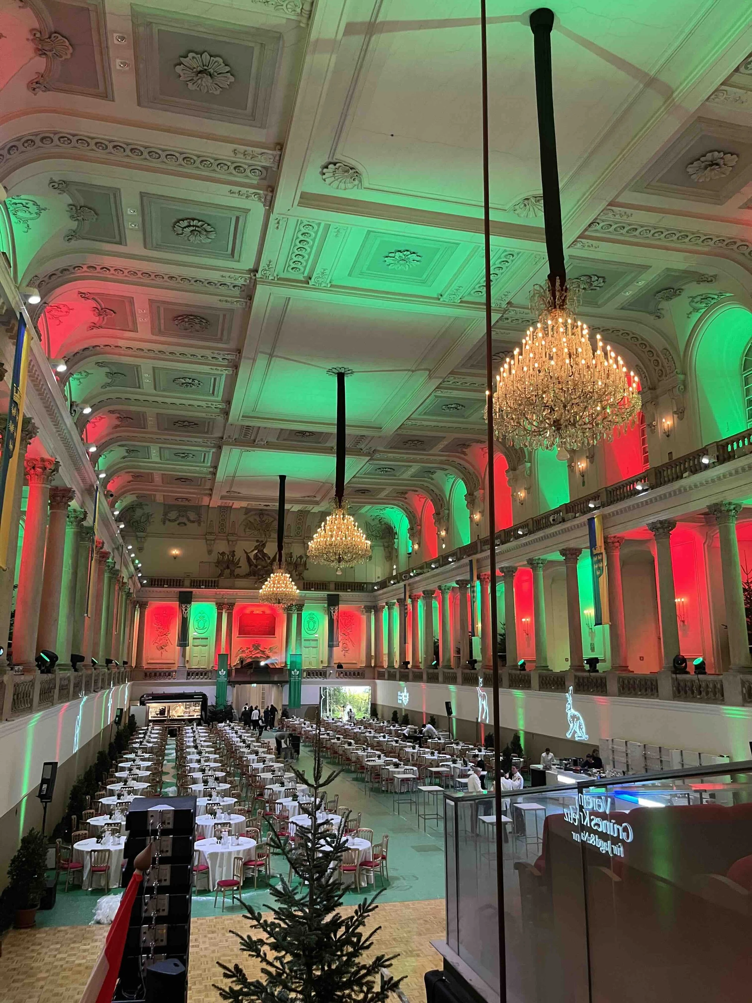 Die spanische Hofreitschule in Wien. Festlich gedeckte Tische mit weißen Tischdecken und Stühlen. Es gibt einige Personen, die den Raum vorbereiten, und einen Bereich mit Bühne und Ständen im vorderen Bereich.