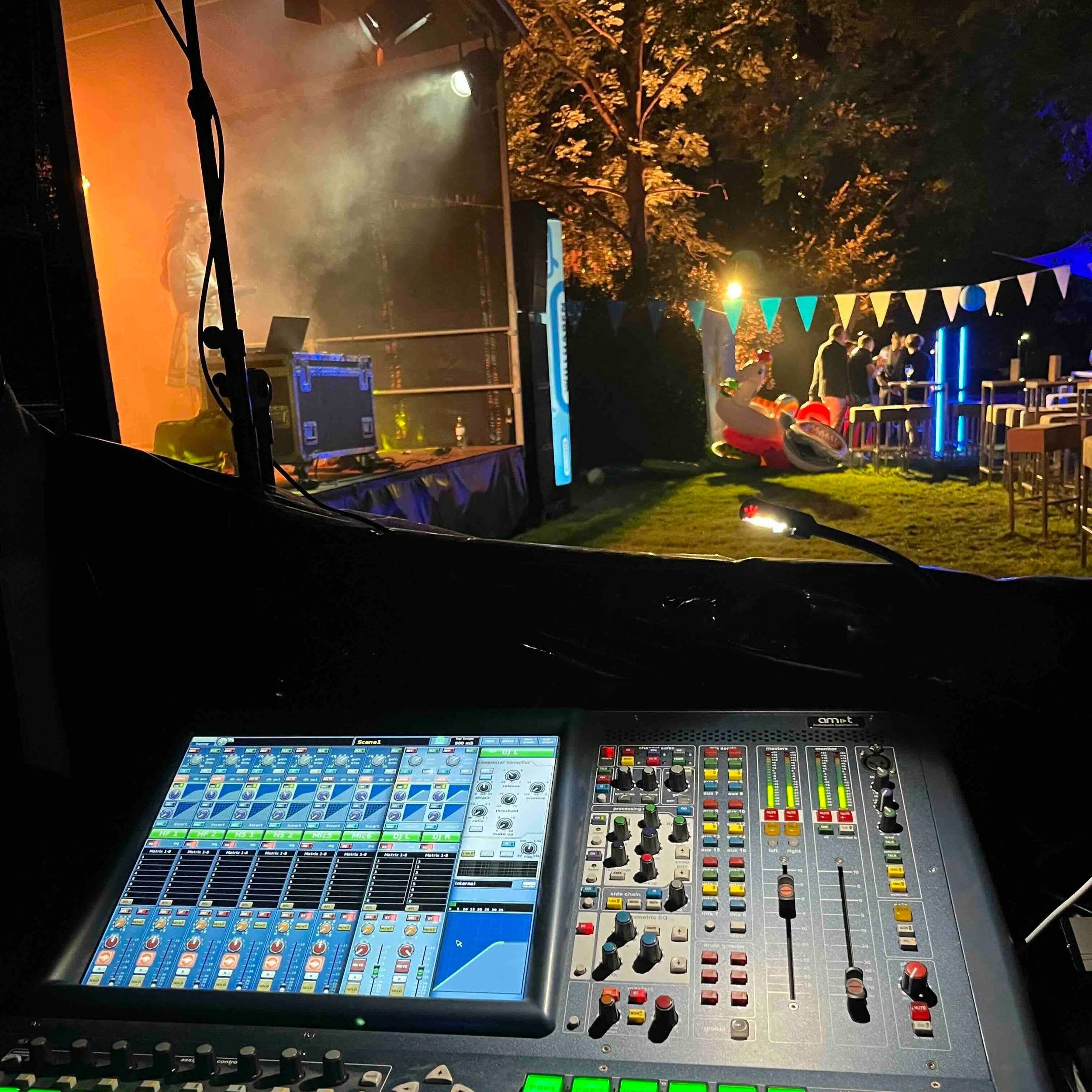 Blick auf das Midas Pro1 Mischpult während einer Veranstaltung im Freien bei Nacht. Im Hintergrund sind eine Gruppe von Menschen, bunte Flaggen und eine aufblasbare Entenfigur sichtbar.