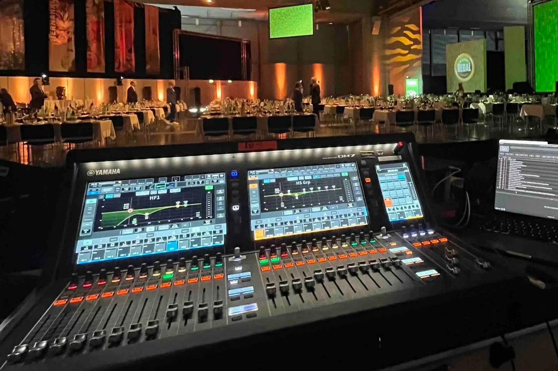 Ein Yamaha DM7 Tonmischpult in einem Veranstaltungsraum, der für eine Feier oder Gala vorbereitet ist. Im Hintergrund sind gedeckte Tische und Stühle, während die Wände des Saals mit bunten Lichtern beleuchtet sind.