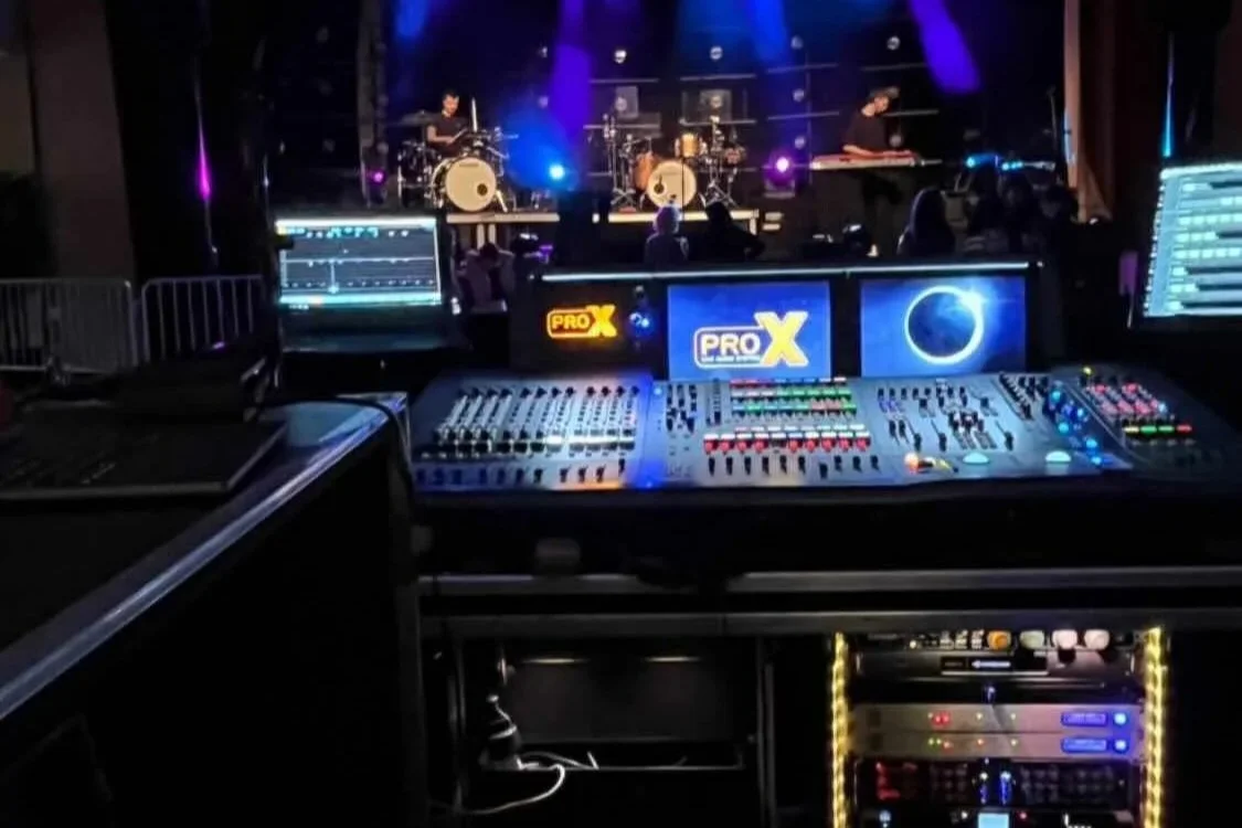 Bühne mit drei Musikern und einem Schlagzeug, farbiges Bühnenlicht, im Vordergrund ein Midas ProX Mischpult und Monitore.