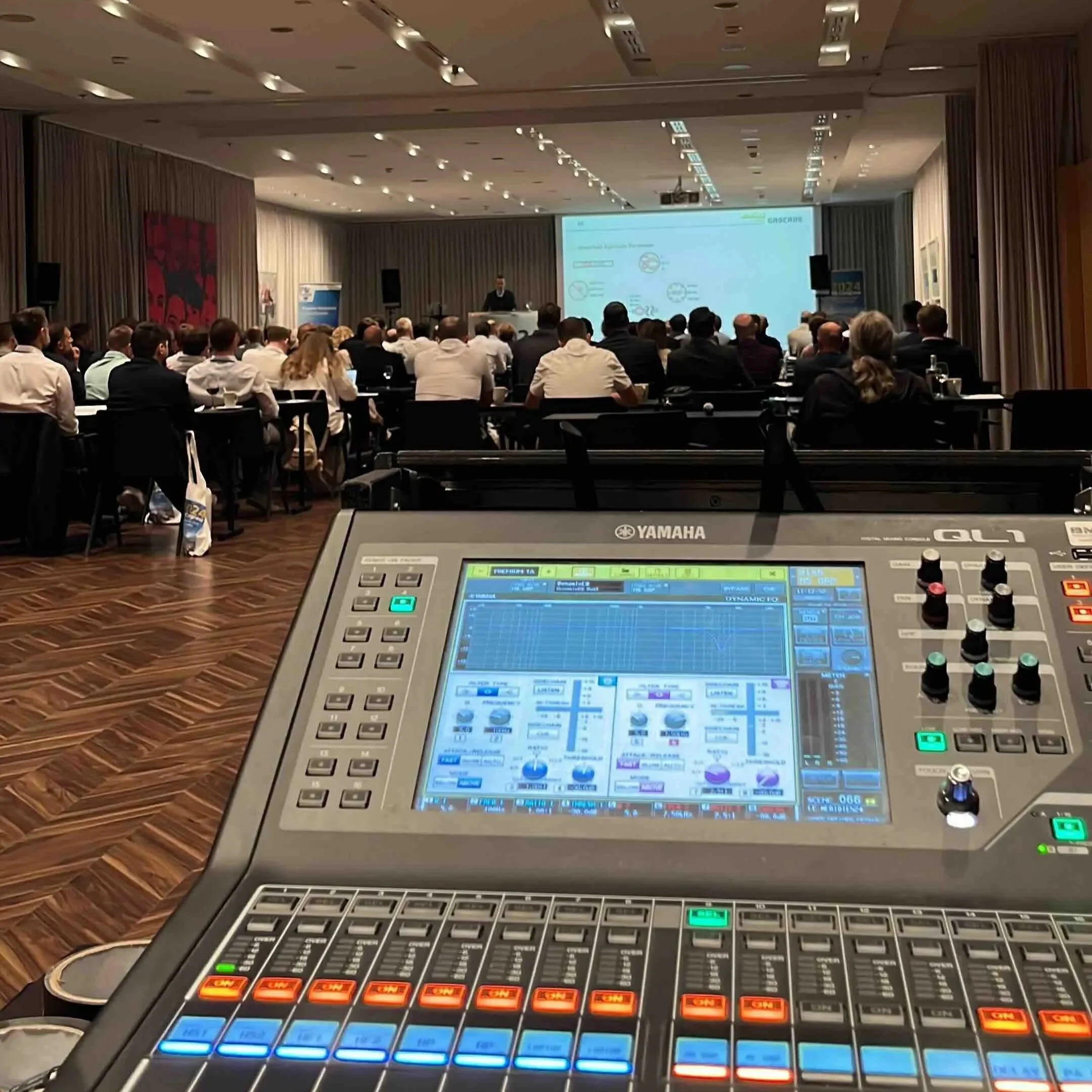 Konferenzraum mit Präsentation auf einer großen Leinwand, zur linken ein Rotes Gemälde mit schwarzem Muster, im Vordergrund ein Yamaha QL1 Tonmischpult, das auf eine Bühne und einen Bereich mit Sitzplätzen blickt.