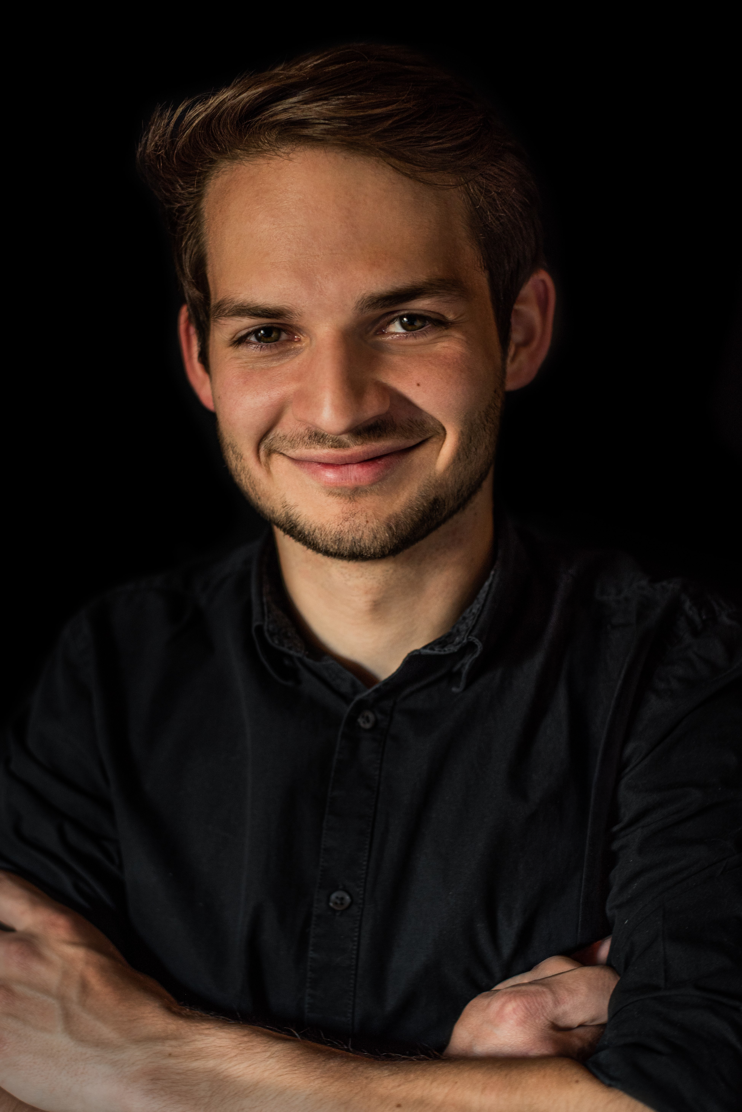 Ein Foto von Michael Gansberger mit braunen Haaren und freundlichem Lächeln, trägt ein schwarzes Hemd vor schwarzem Hintergrund.