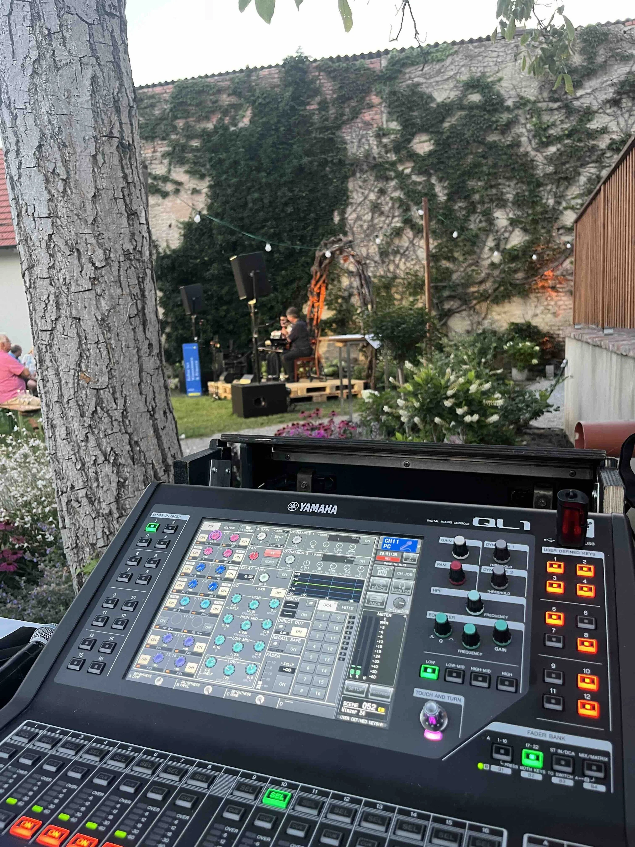 Ein Yamaha QL1 Mischpult auf einer Bühne während eines Live-Musikauftritts im Freien mit einem Musiker im Hintergrund, umgeben von Pflanzen und Dekorationen.