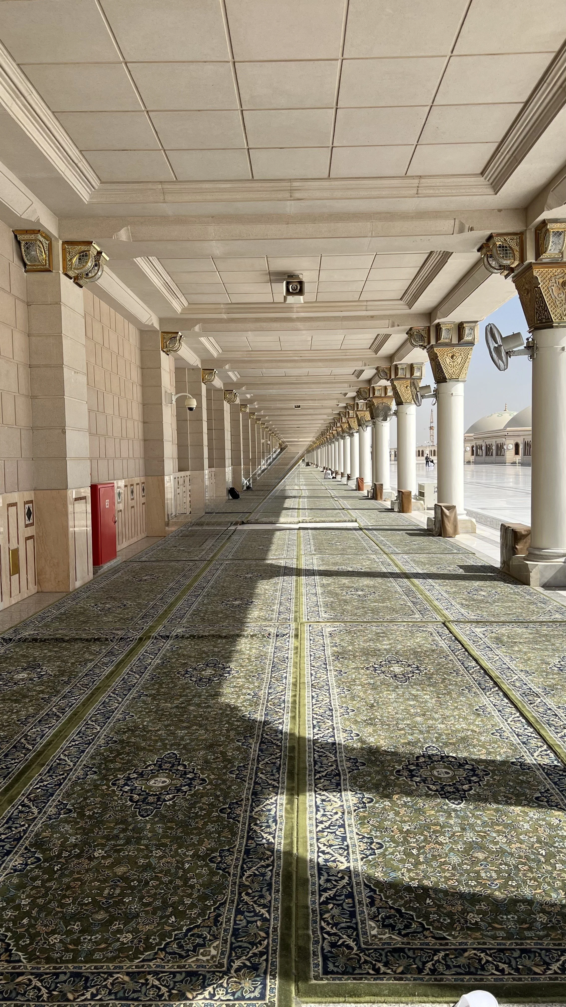 vue d'un long couloir avec tapisseries orientales, des colonnes blanches ornées de dorures, et un ciel dégagé visible à l'extérieur.