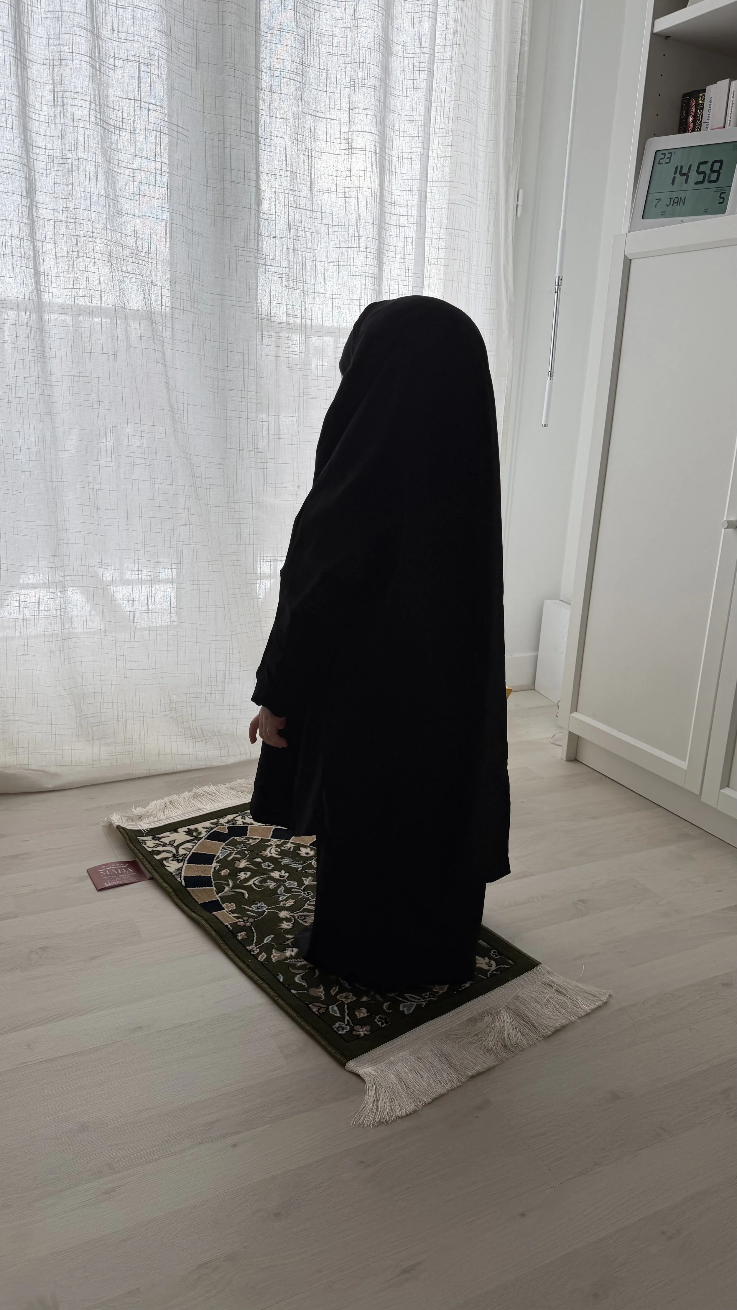 Une personne en tenues traditionnelles musulmanes priant sur un tapis de prière dans une pièce lumineuse avec des rideaux blancs