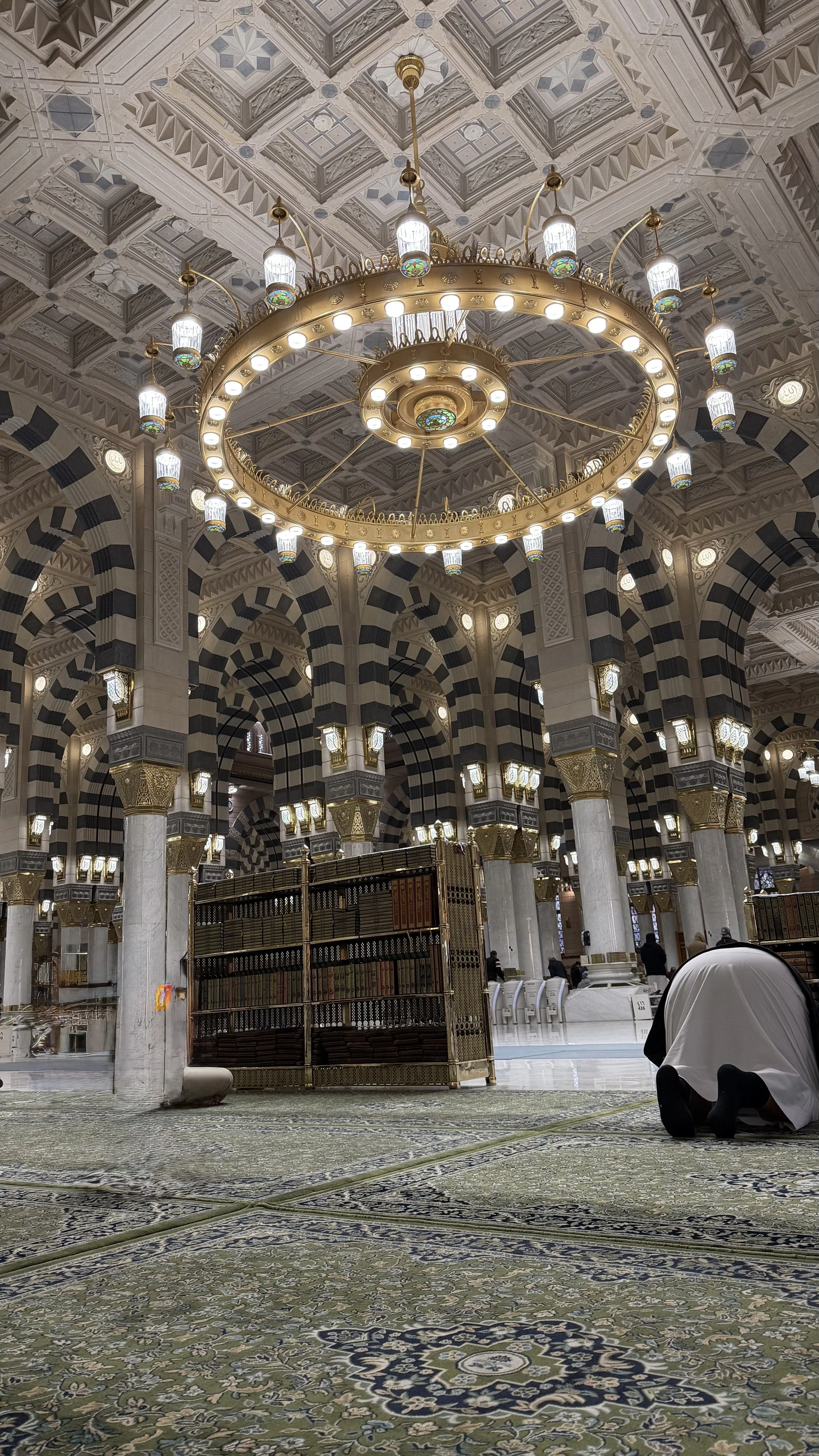 Intérieur d'une mosquée avec un plafond orné, une grande chandelier circulaire suspendu, des arches noires et blanches, et des personnes en prière sur un tapis complexe.