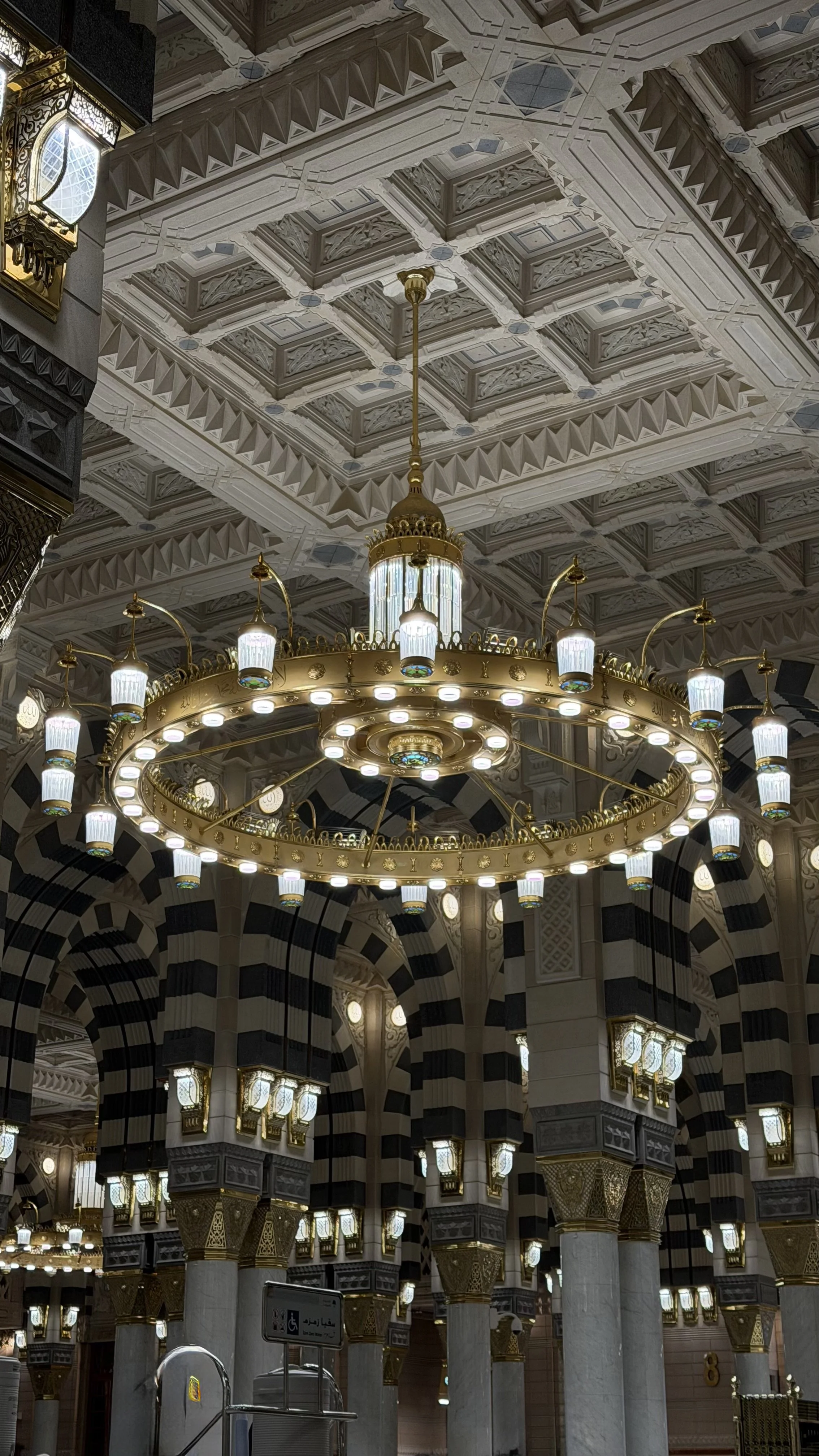 Un plafond orné d'un grand lustre circulaire en or avec des lampes blanches suspendues dans une mosquée, entouré de colonnes décorées avec des motifs en or et noir et des murs avec des arcades et des lumières. 