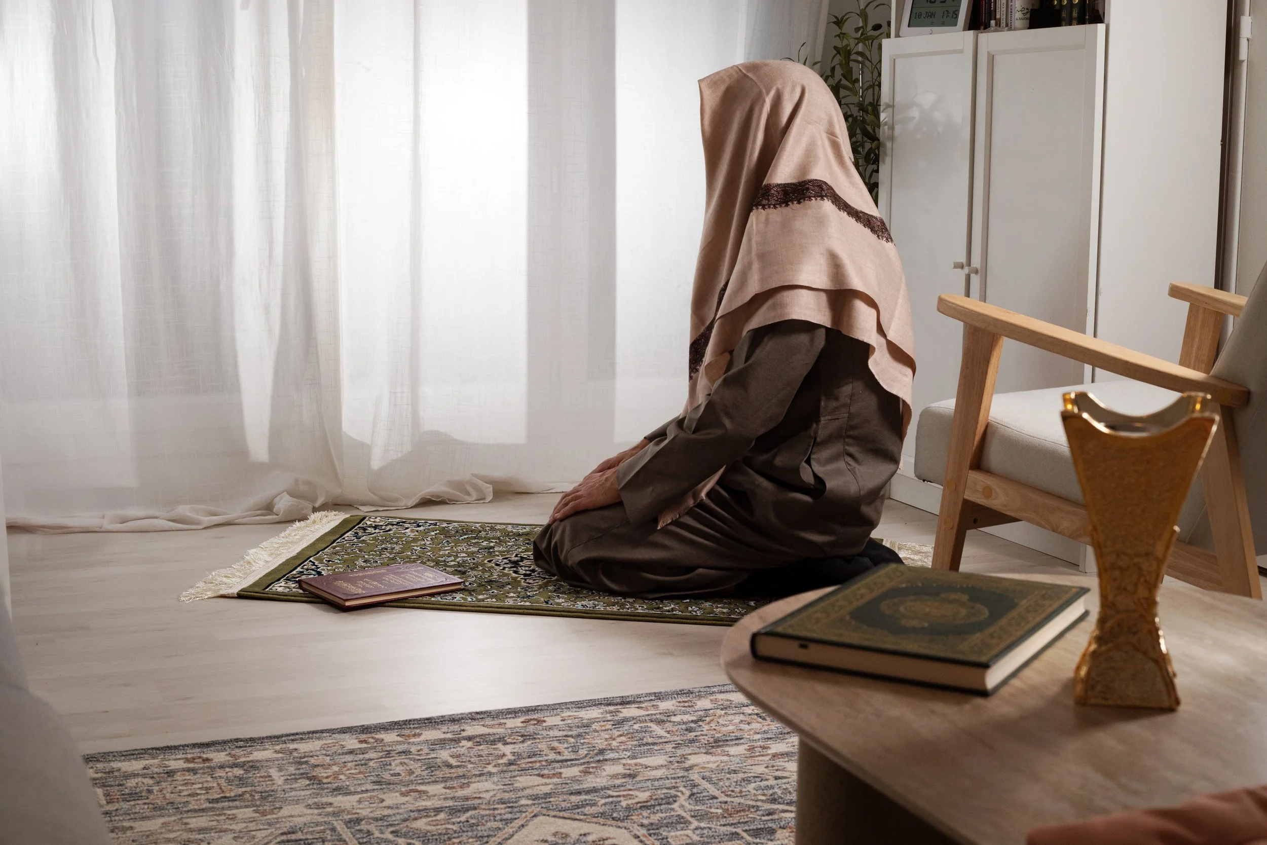 Une femme en hijab priante à genoux dans une pièce lumineuse avec rideaux blancs, un tapis vert, et des livres sur une table en bois.