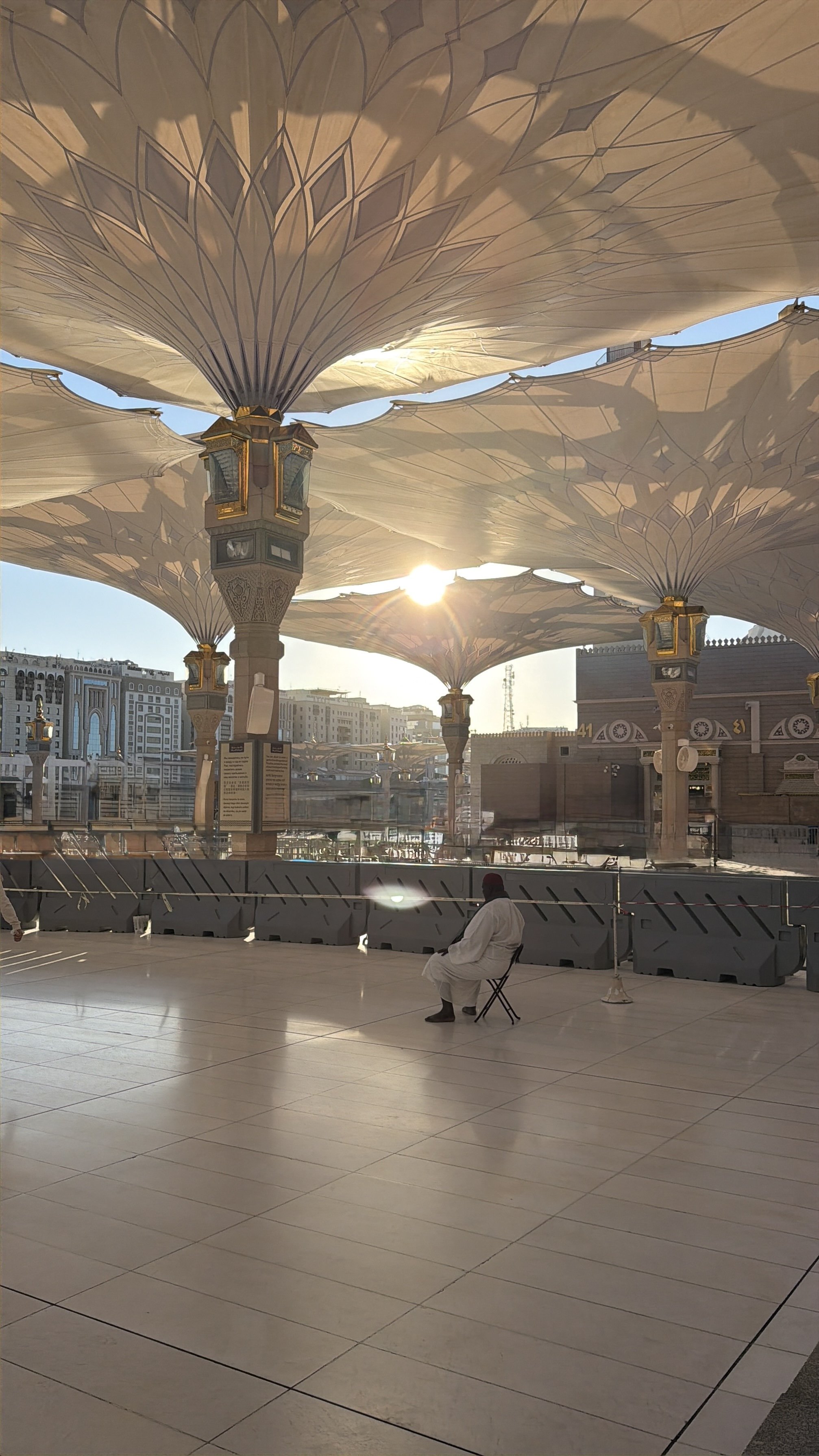 Personne assise sous un grand toit décoratif en forme de fleurs avec le soleil couchant en arrière-plan, dans un lieu apparenté à une mosquée ou une place publique en architecture islamique.