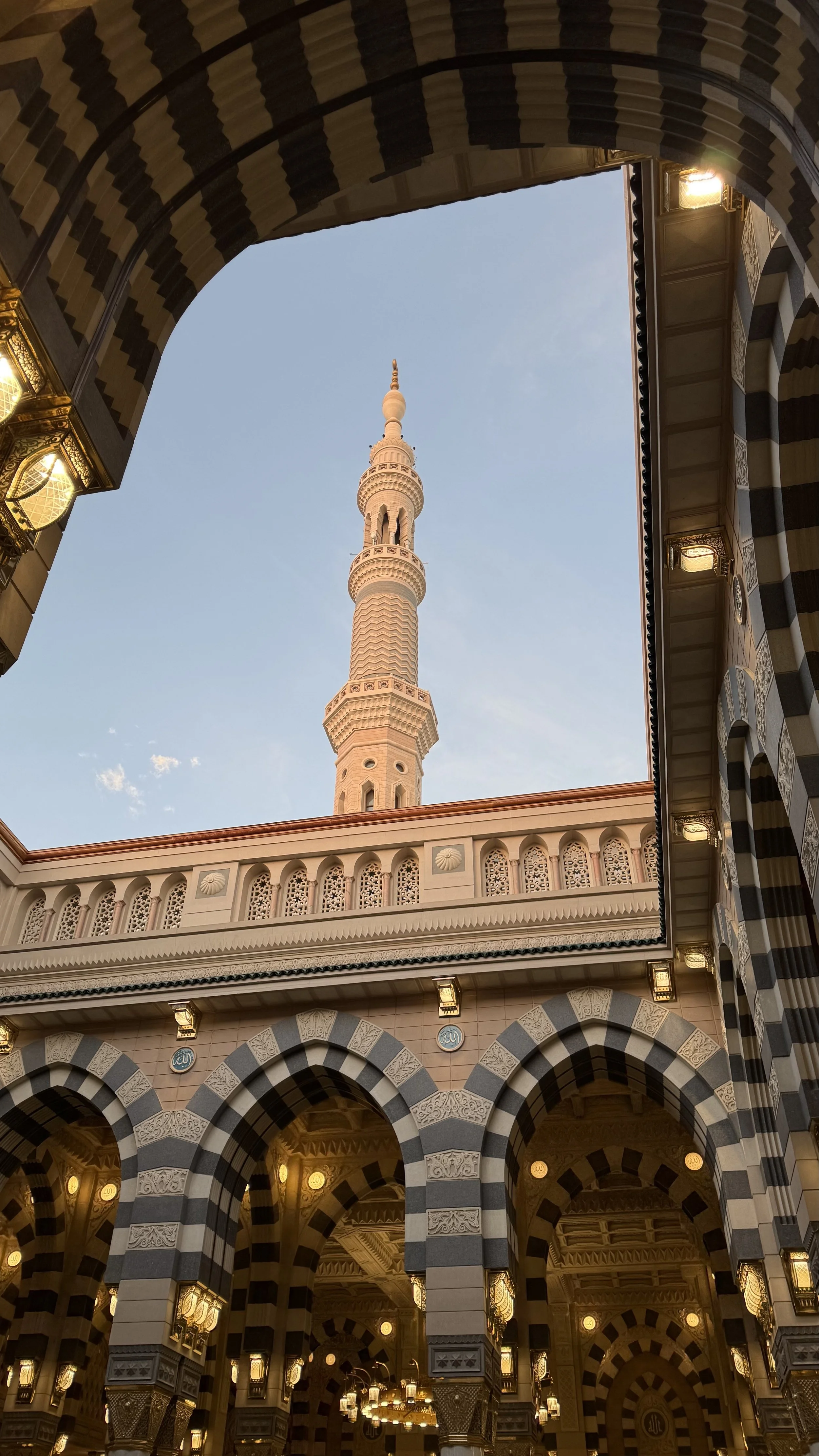 Vue d'une mosquée avec un minaret visible par une ouverture dans la cour, structure en arches avec motifs en noir et blanc, lumières suspendues à l'intérieur.