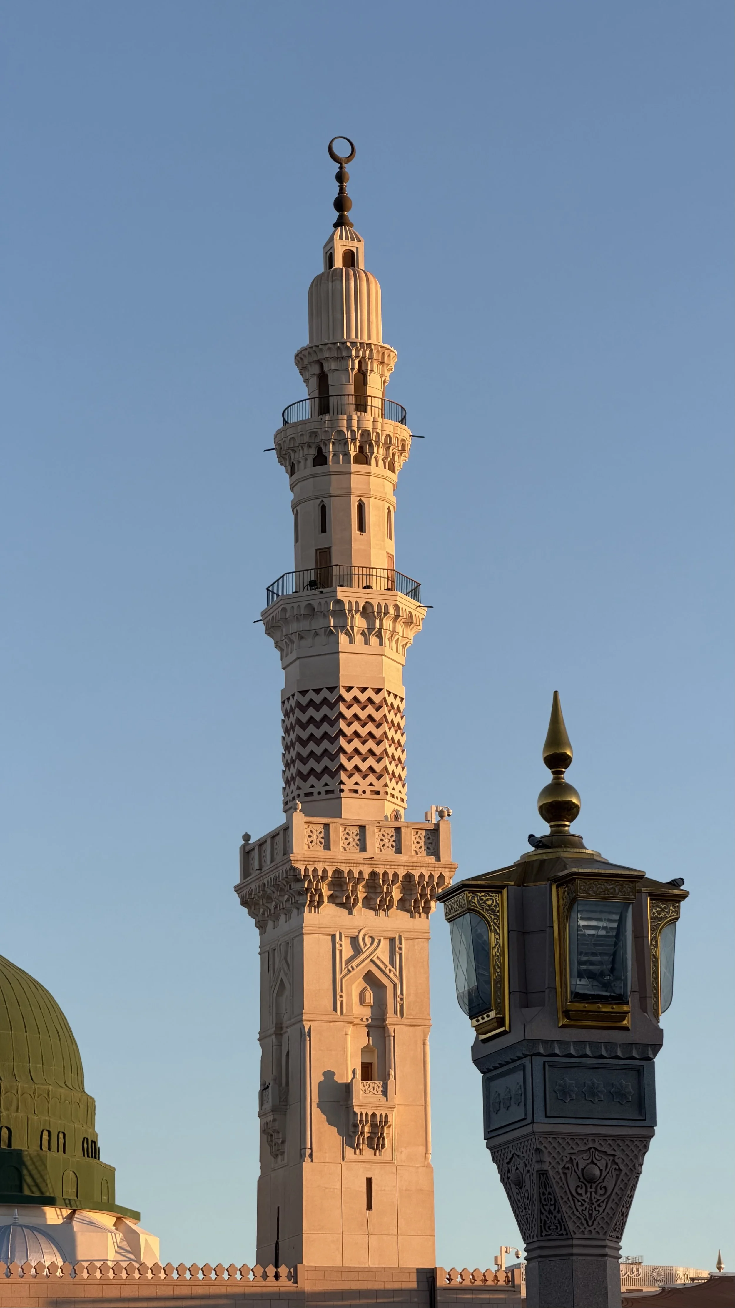 Une tour de minaret de couleur beige claire avec plusieurs niveaux décorés, sous un ciel bleu clair, avec en premier plan un lampadaire noir avec des détails dorés.